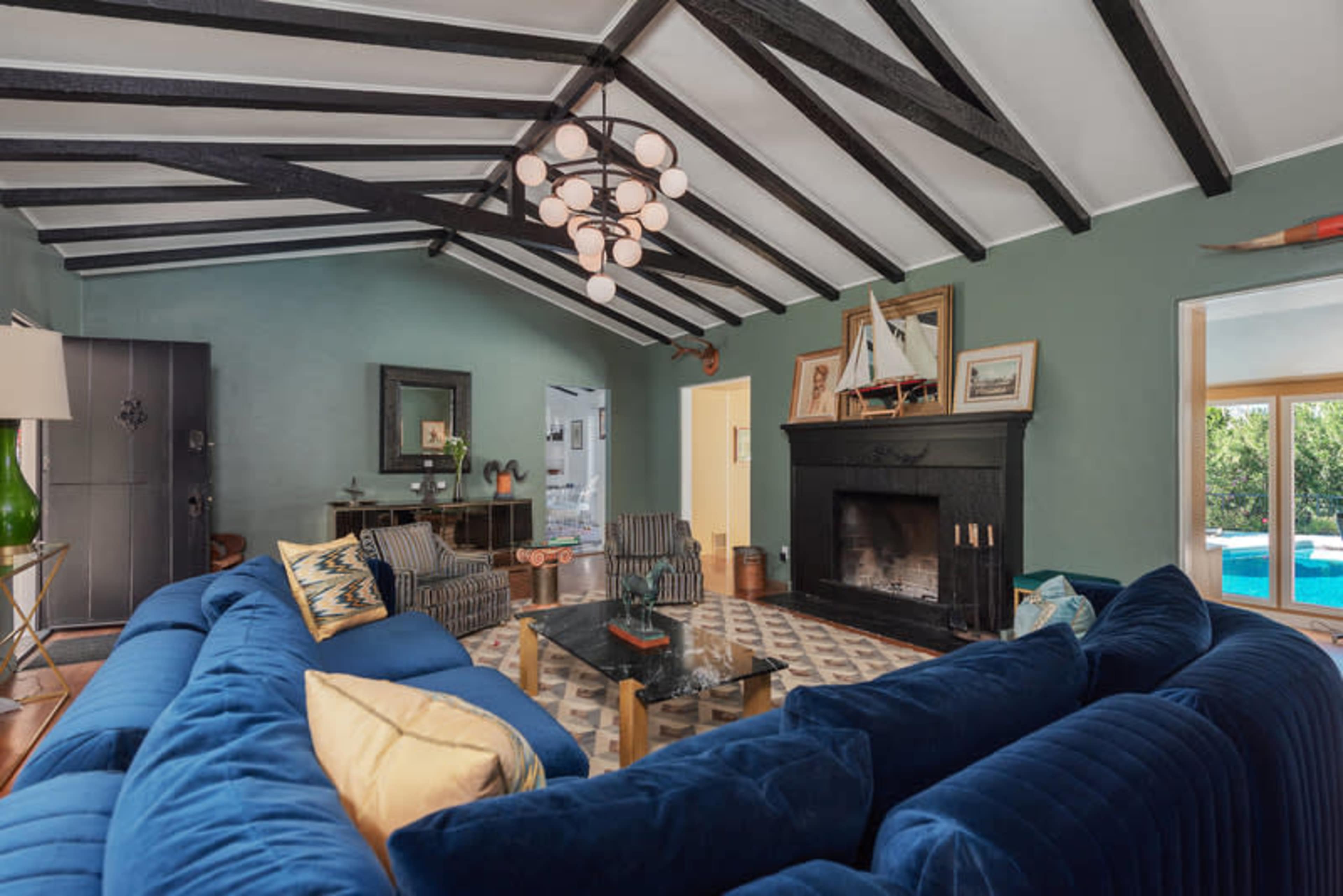 The living room features a blue sectional sofa, a glass coffee table, and a fireplace, all set under exposed wooden beams.