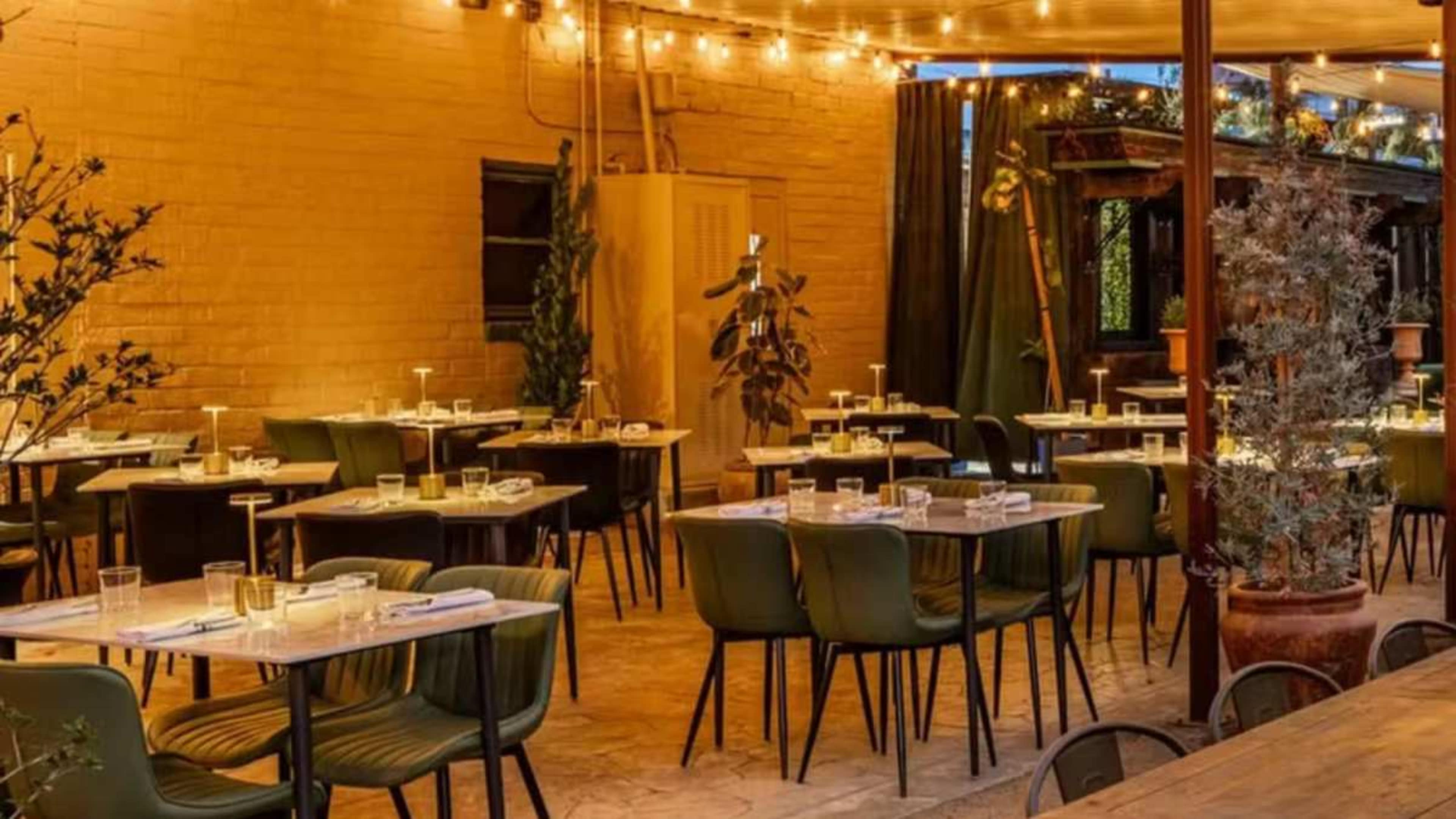 The image shows an outdoor dining area of a restaurant with tables set for patrons, surrounded by plants and string lights.