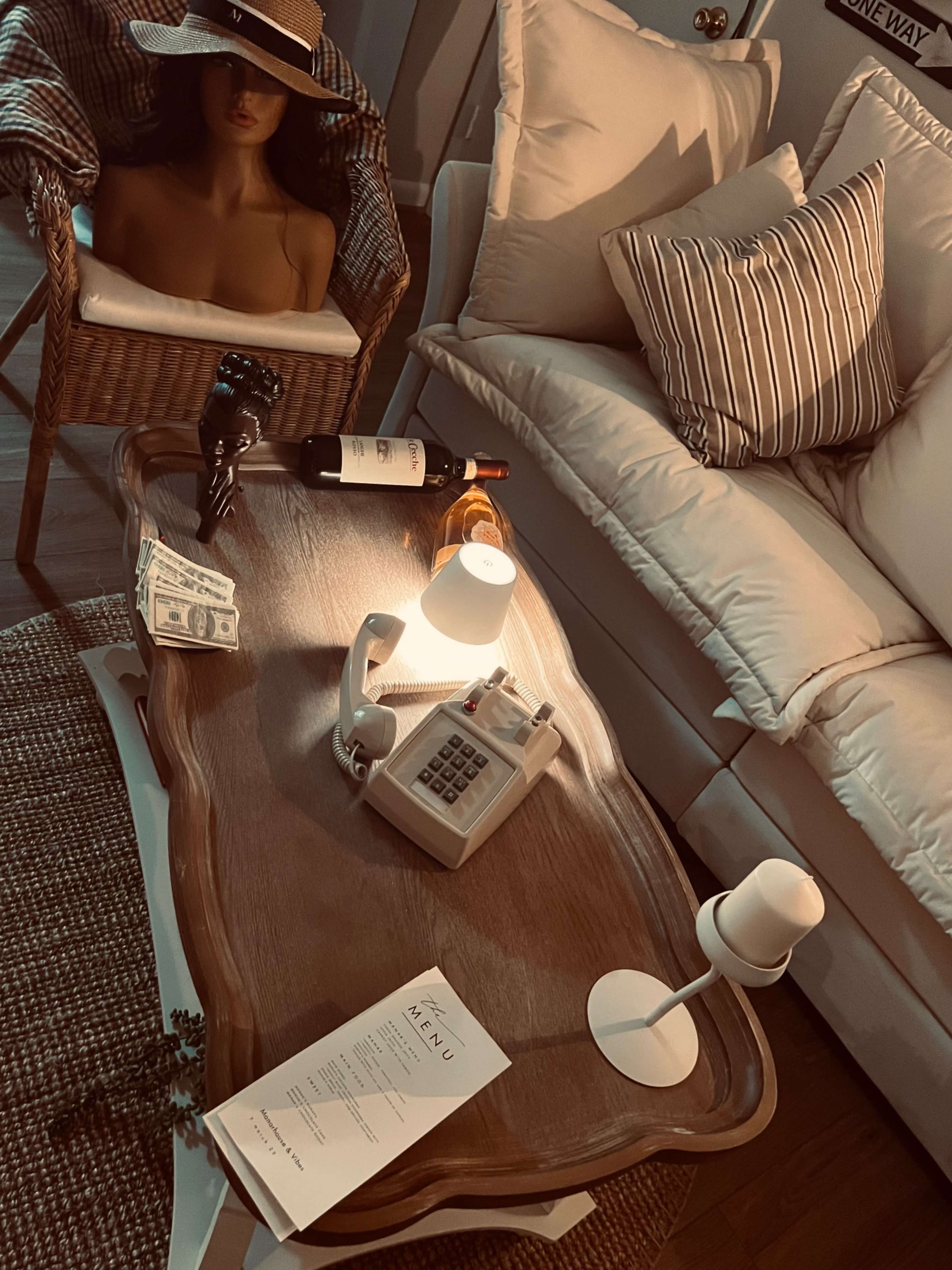The scene features a coffee table with a wine bottle, cash, an old-fashioned telephone, a menu, and a table lamp, surrounded by a wicker chair and a sofa with cushions.