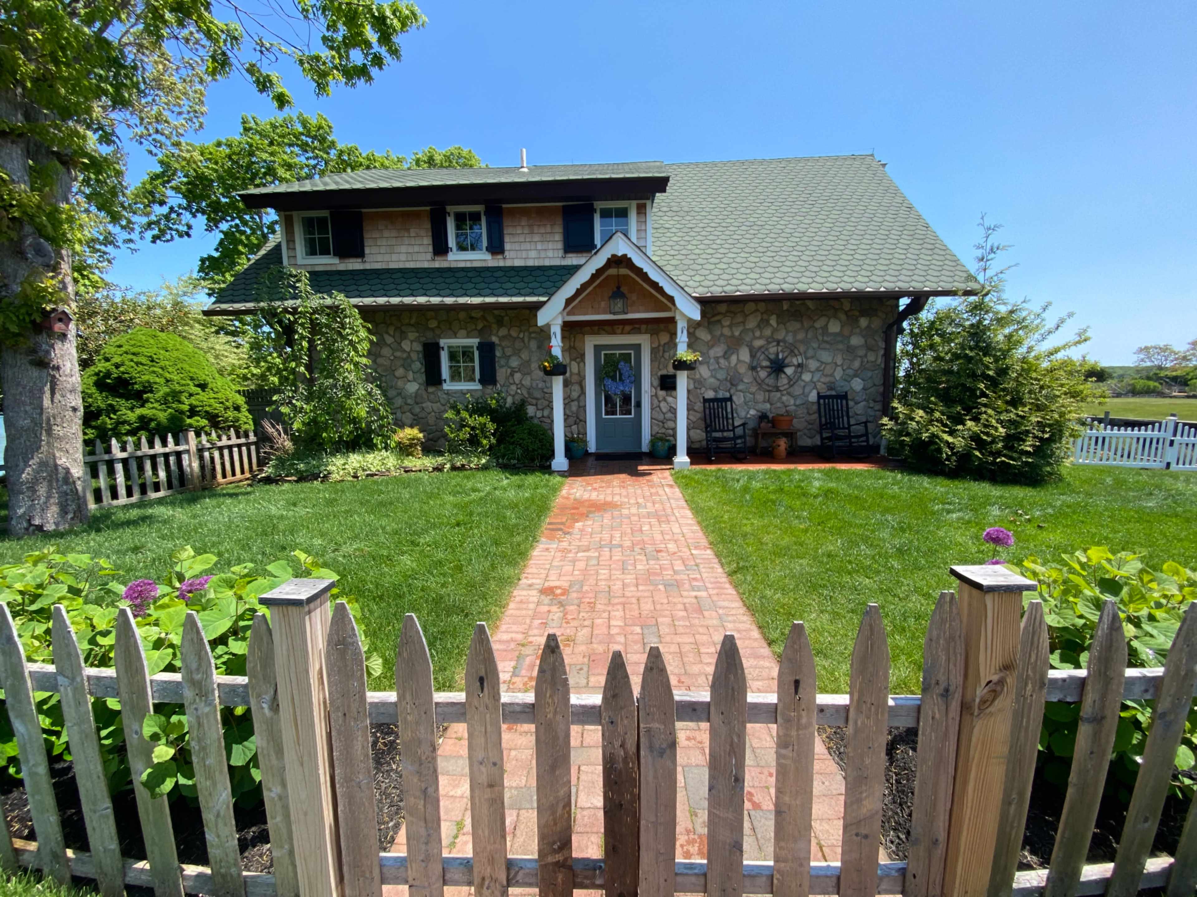 A two-story stone house with a green roof and a brick walkway, surrounded by grass and garden plants, is set behind a wooden picket fence.