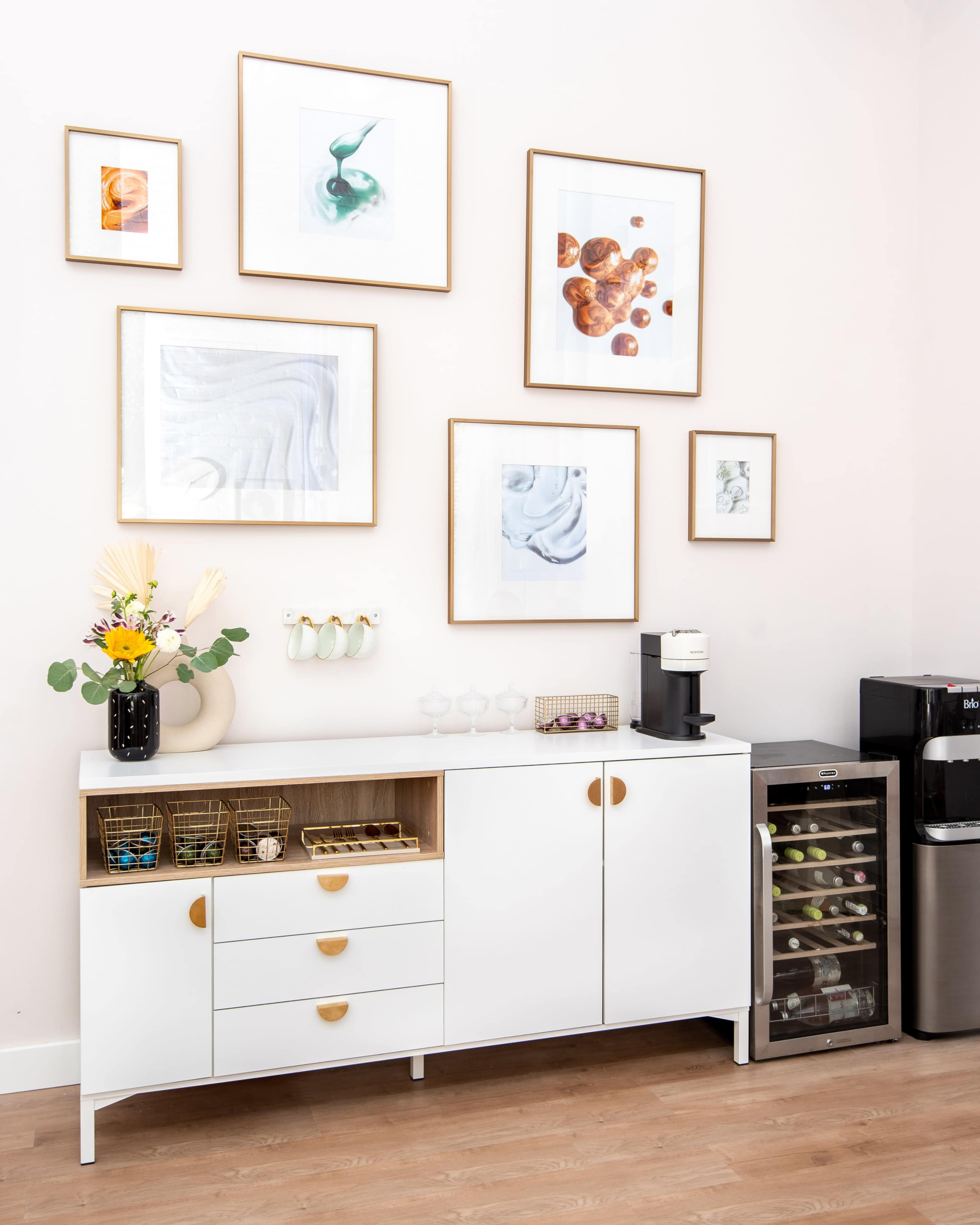 A modern white cabinet with gold handles, adorned with framed artwork on the wall above and a coffee maker on one side.