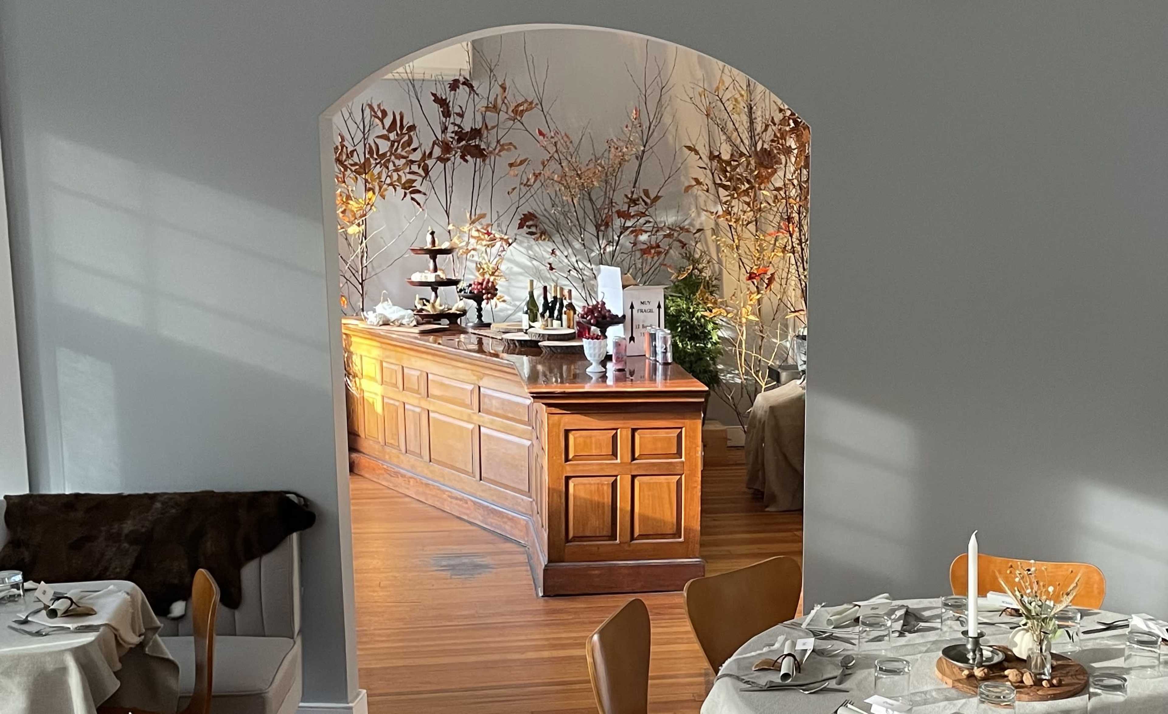 A restaurant interior with a wooden bar, elegantly arranged tables, and decorative branches in the background.