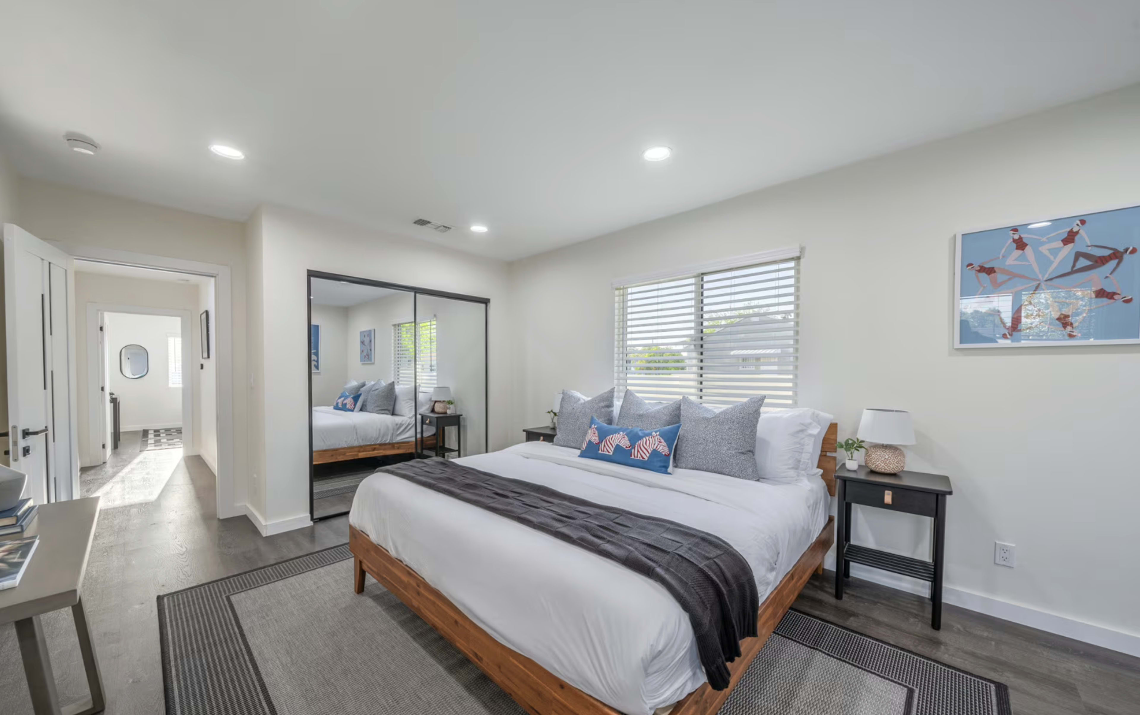 The image shows a modern bedroom featuring a large bed with decorative pillows, a mirror, and a view into a hallway with a patterned floor.