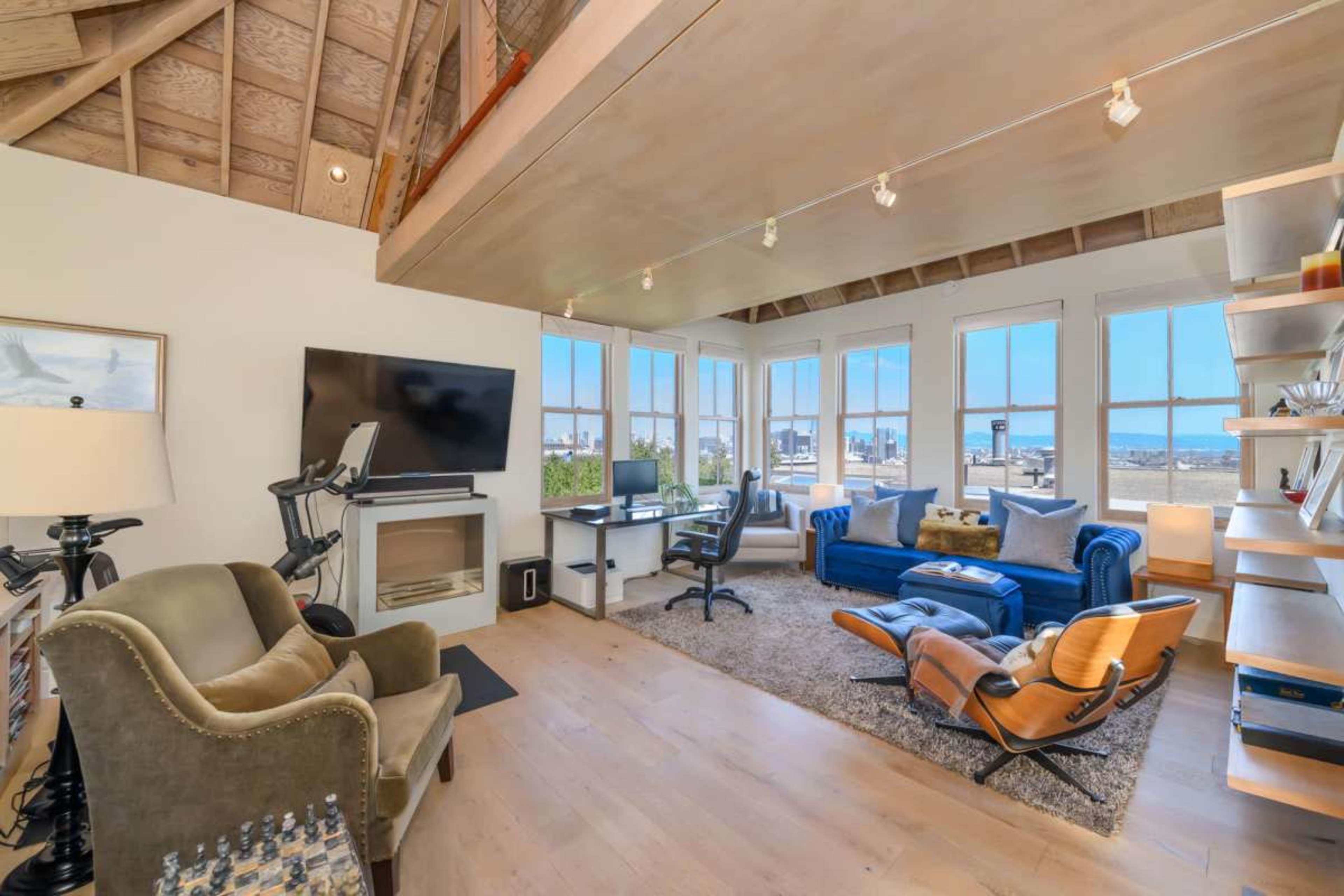 The image depicts a modern living room with a blue couch, a desk, an exercise bike, and large windows offering a view of an outdoor space.
