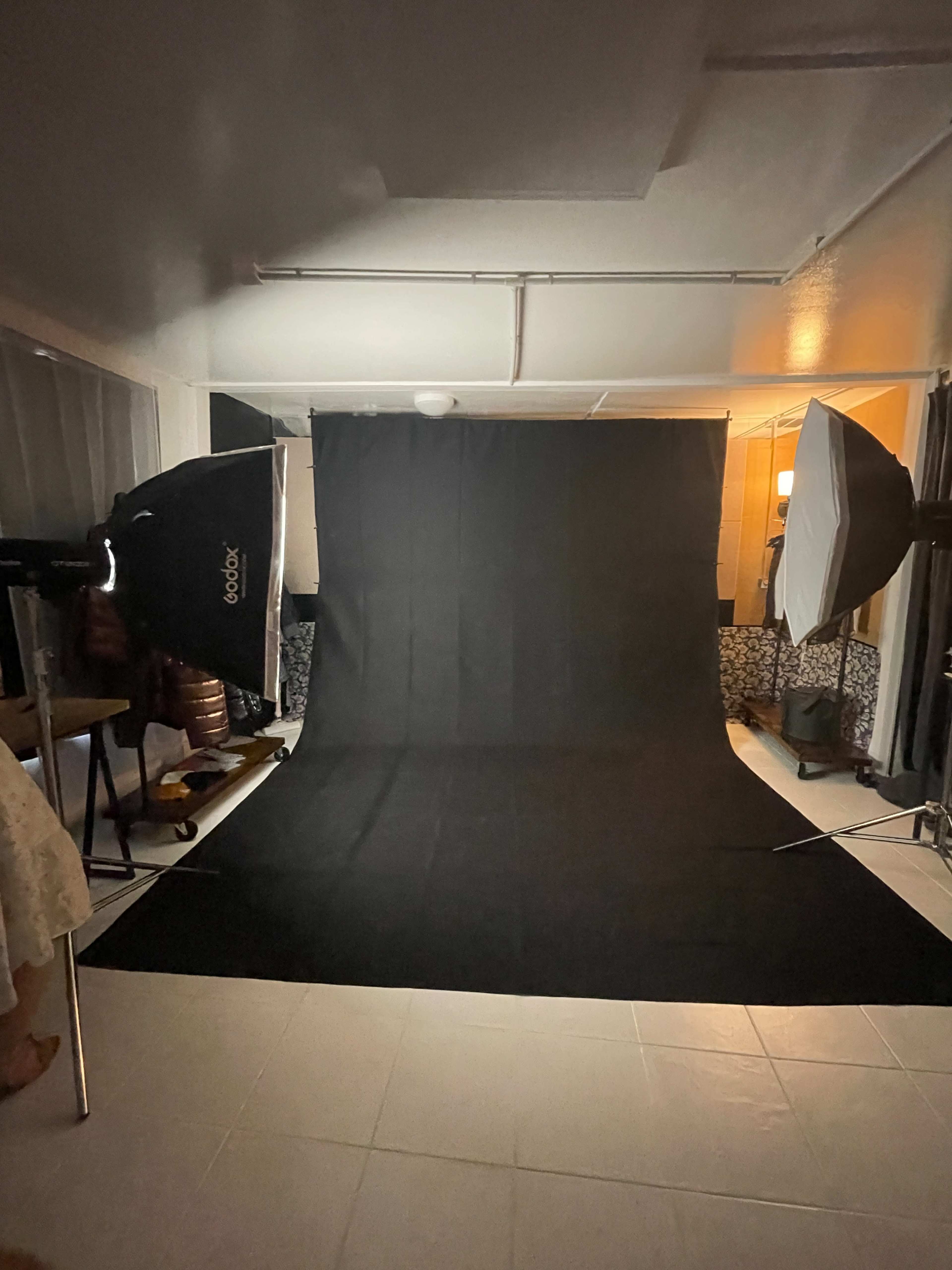 The image shows a photography studio setup with a black backdrop, two softbox lights on either side, and a tiled floor.