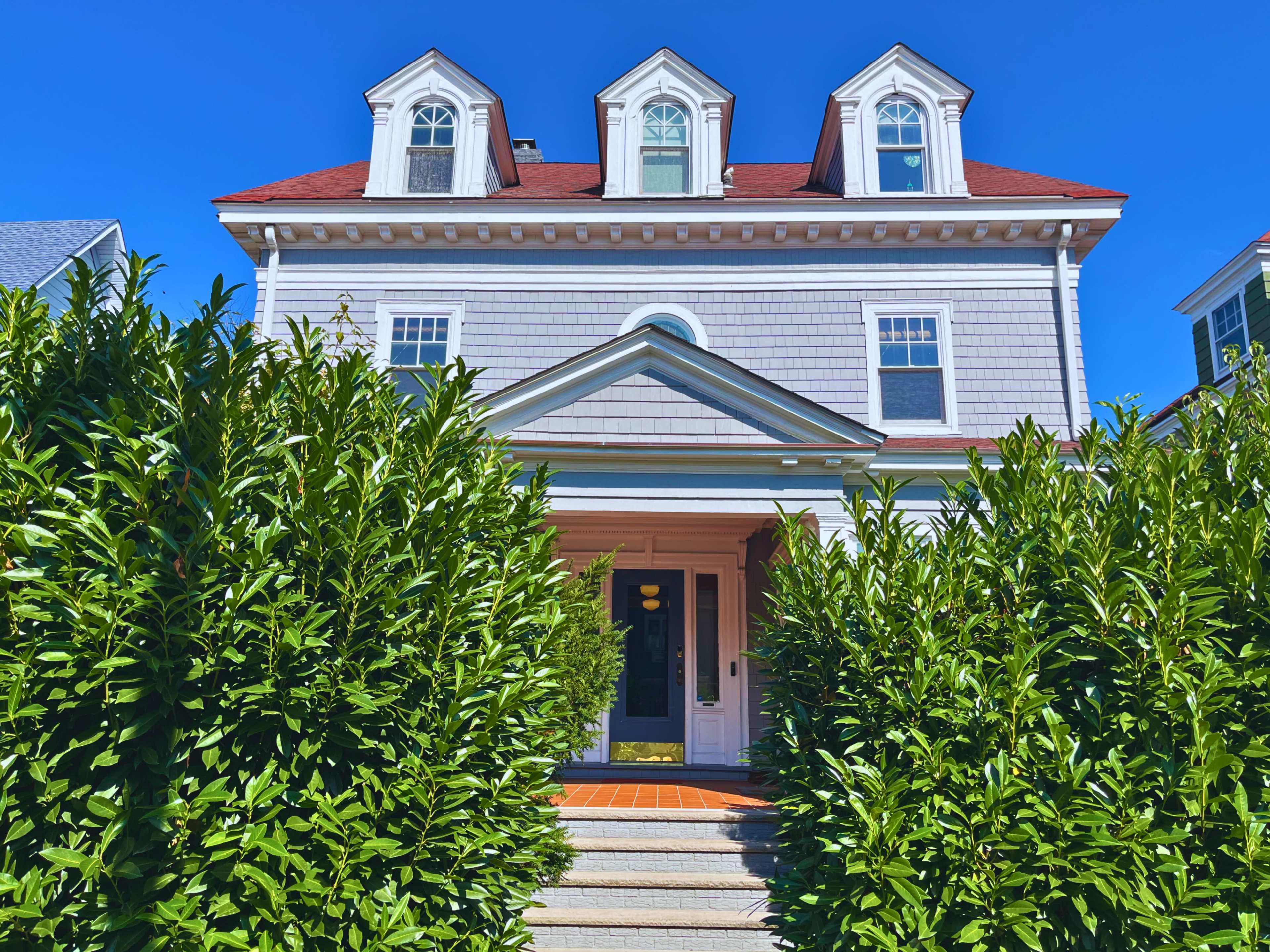 A large gray house with a red roof and three dormer windows is framed by dense green hedges and features a set of stone steps leading to the front door.