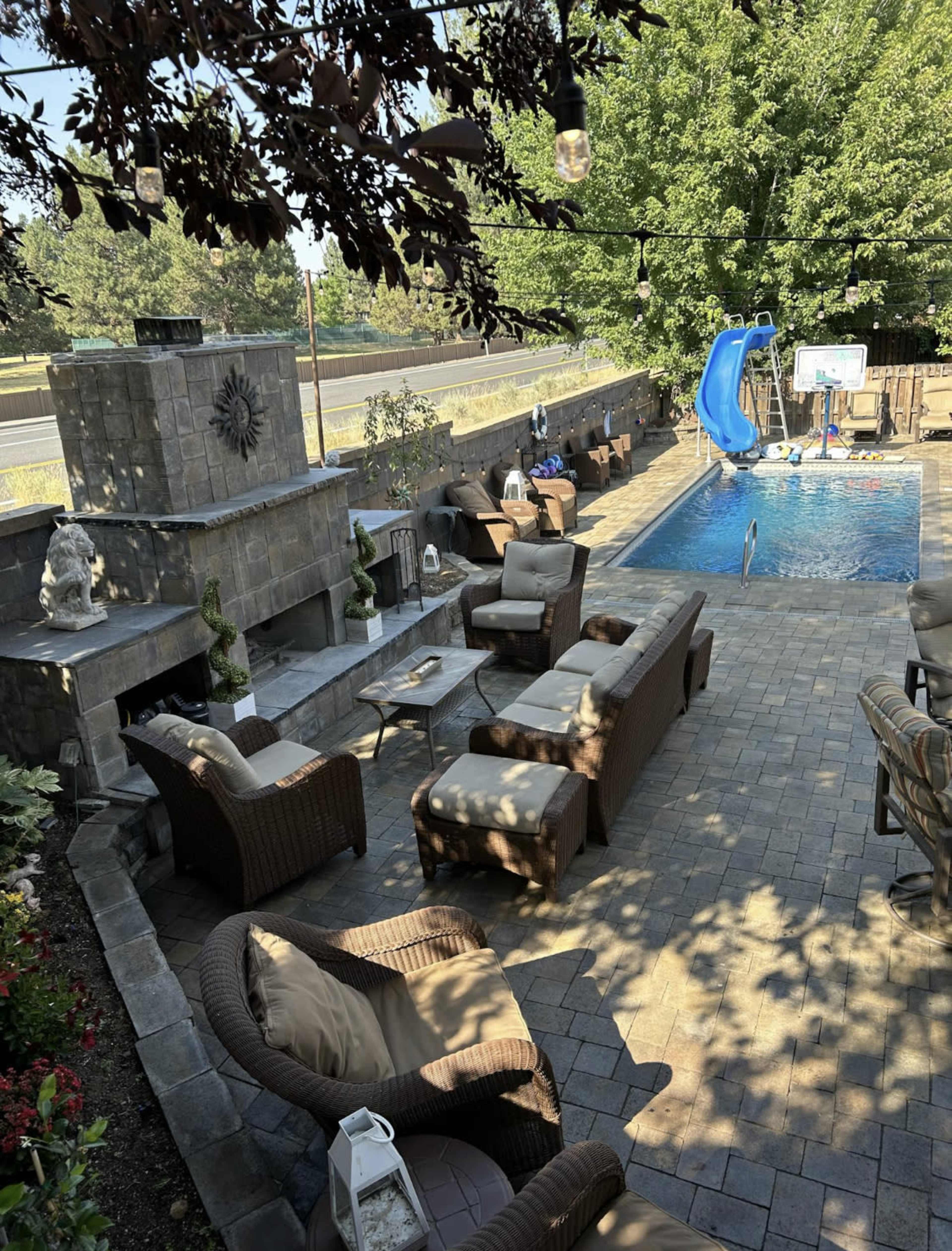 A backyard setting with a swimming pool, a slide, a lounging area with wicker furniture, and a basketball hoop beside the pool.