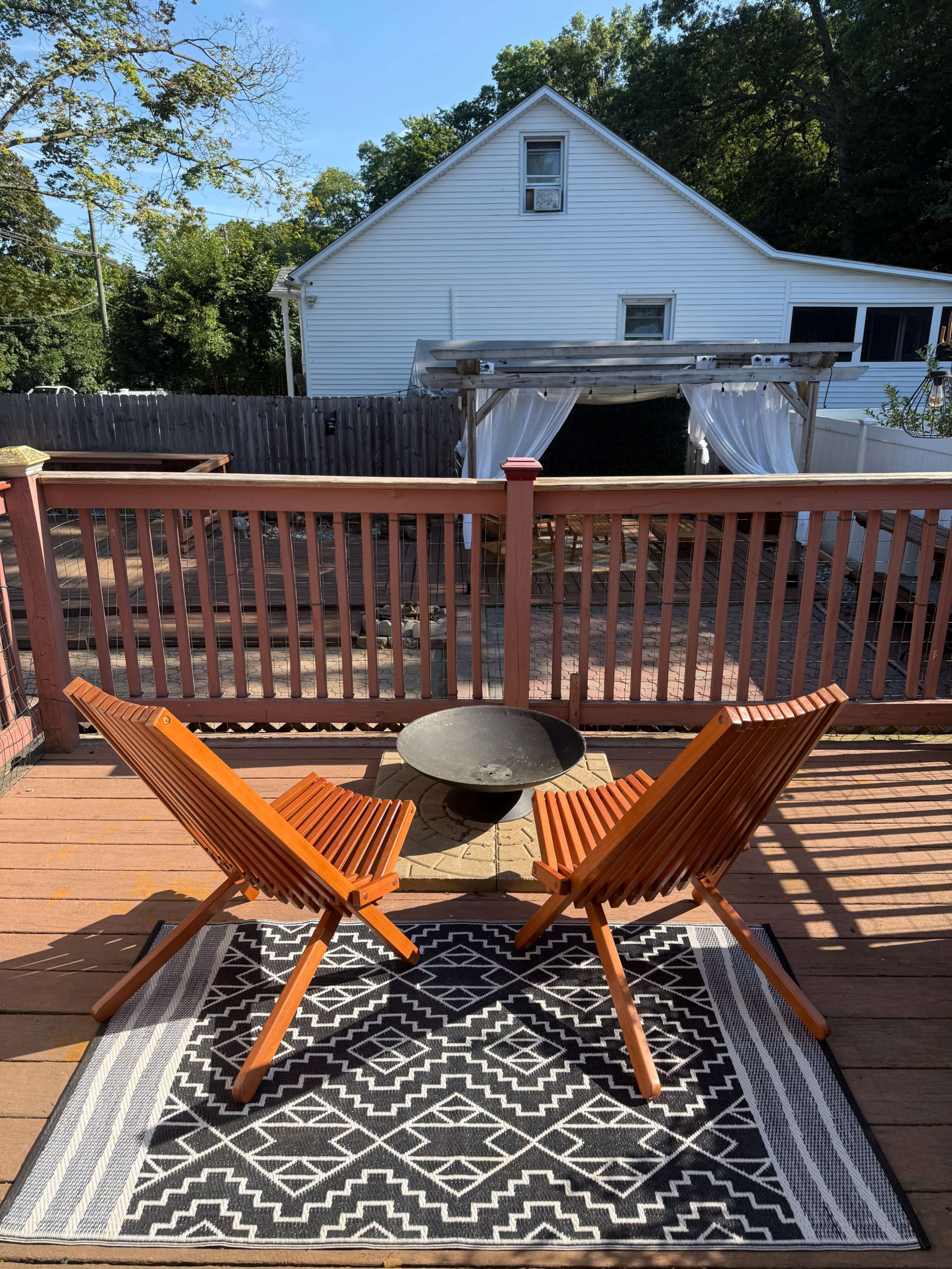 Two wooden chairs face a small fire pit on a deck, with a geometric rug underneath and a house visible in the background.