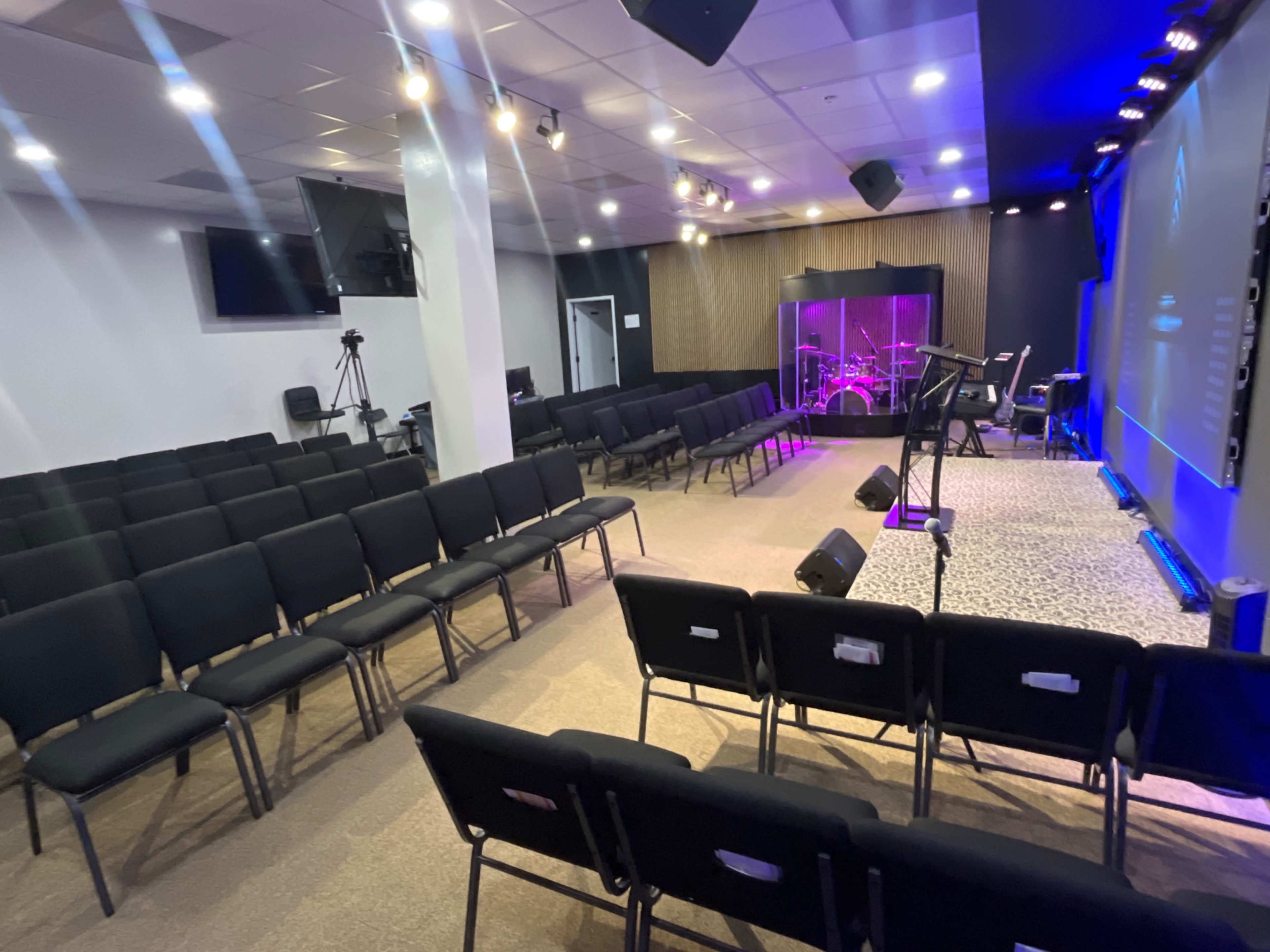 The image shows a modern indoor auditorium with rows of black chairs facing a stage equipped with musical instruments and a screen.