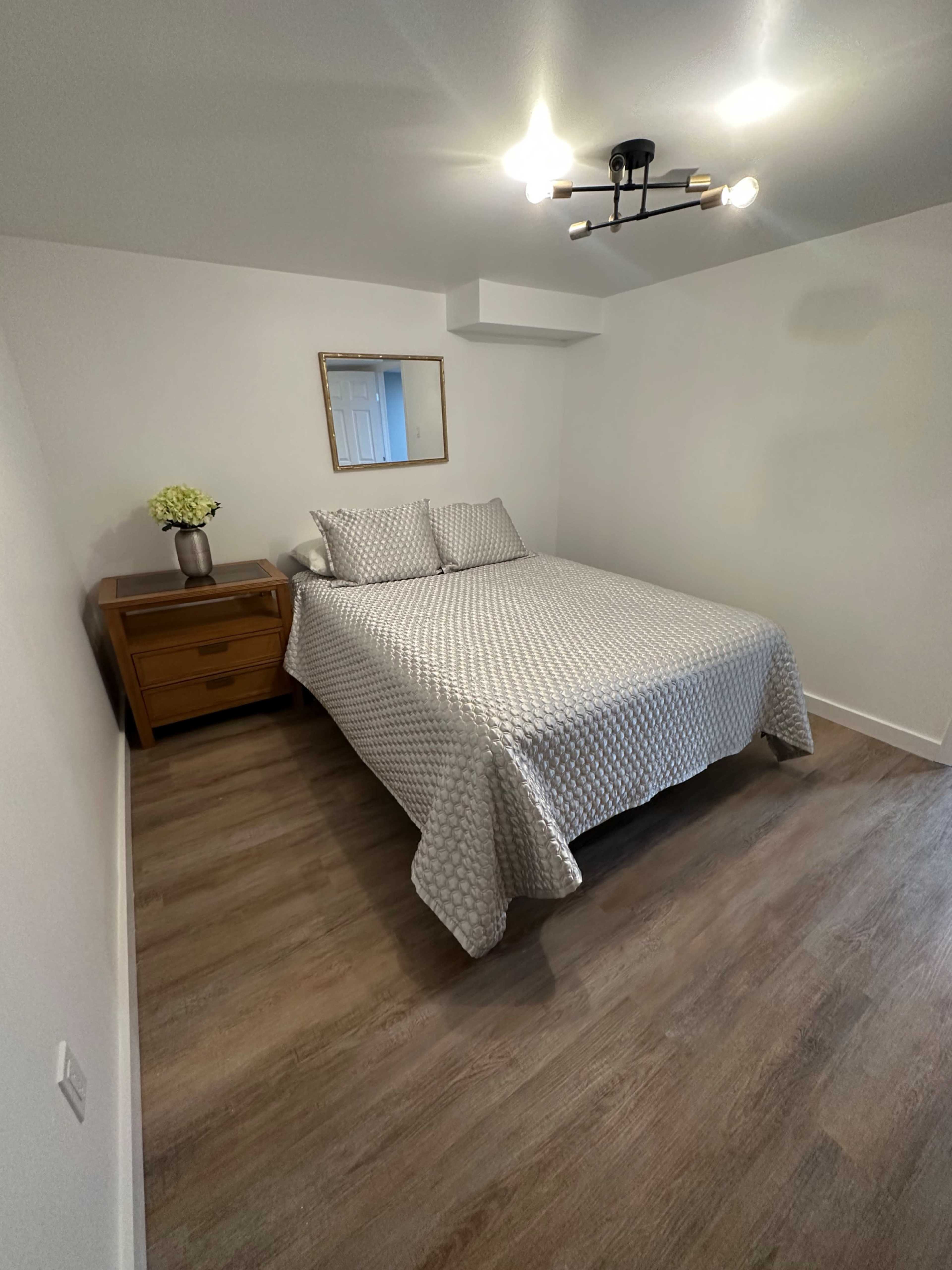 The image shows a simple bedroom with a bed covered in a patterned gray comforter, a wooden nightstand, and a mirror on the wall.