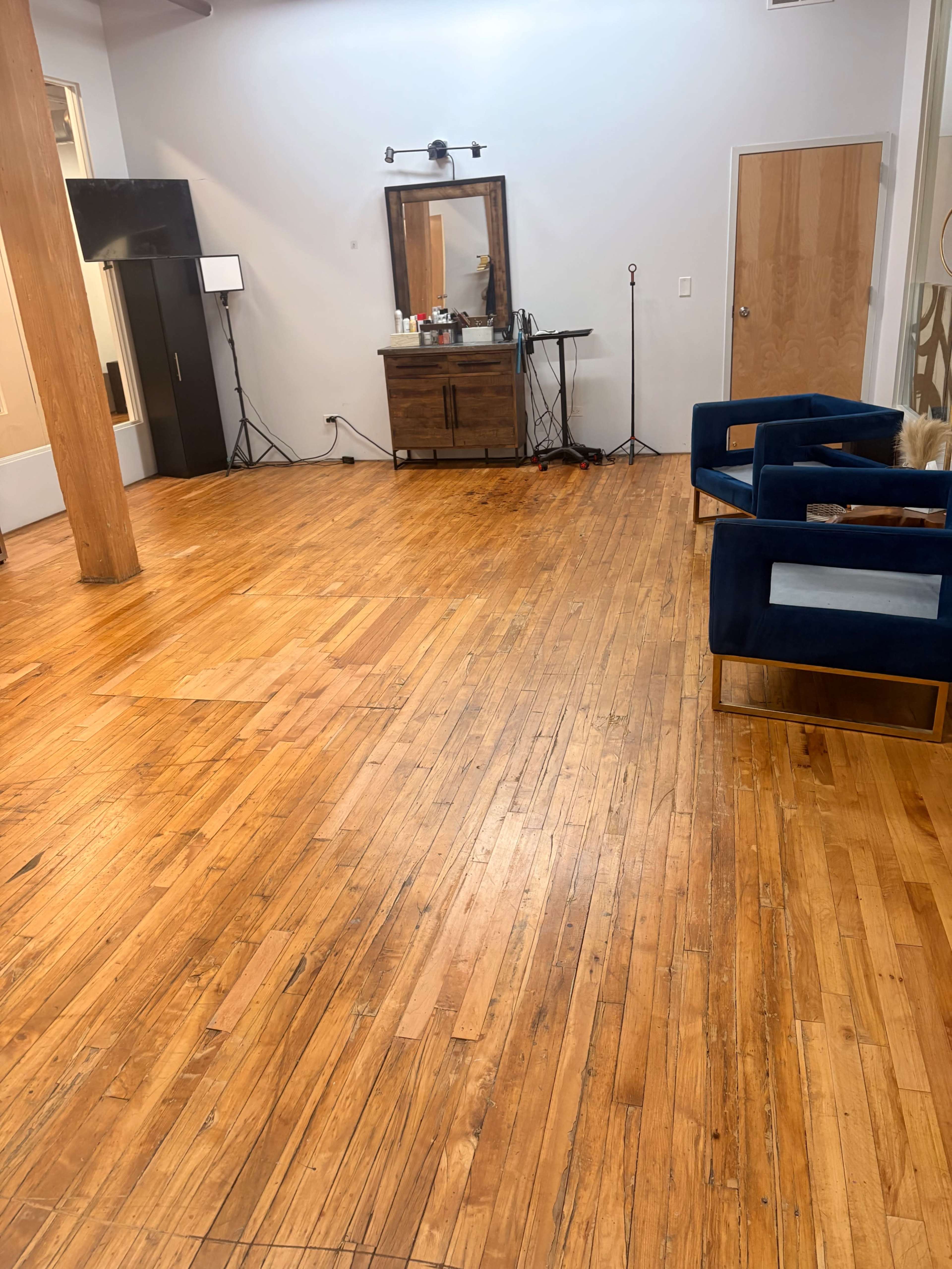 The image shows a spacious, empty room with polished wooden floors, a large mirror, a dresser, and two blue upholstered chairs.