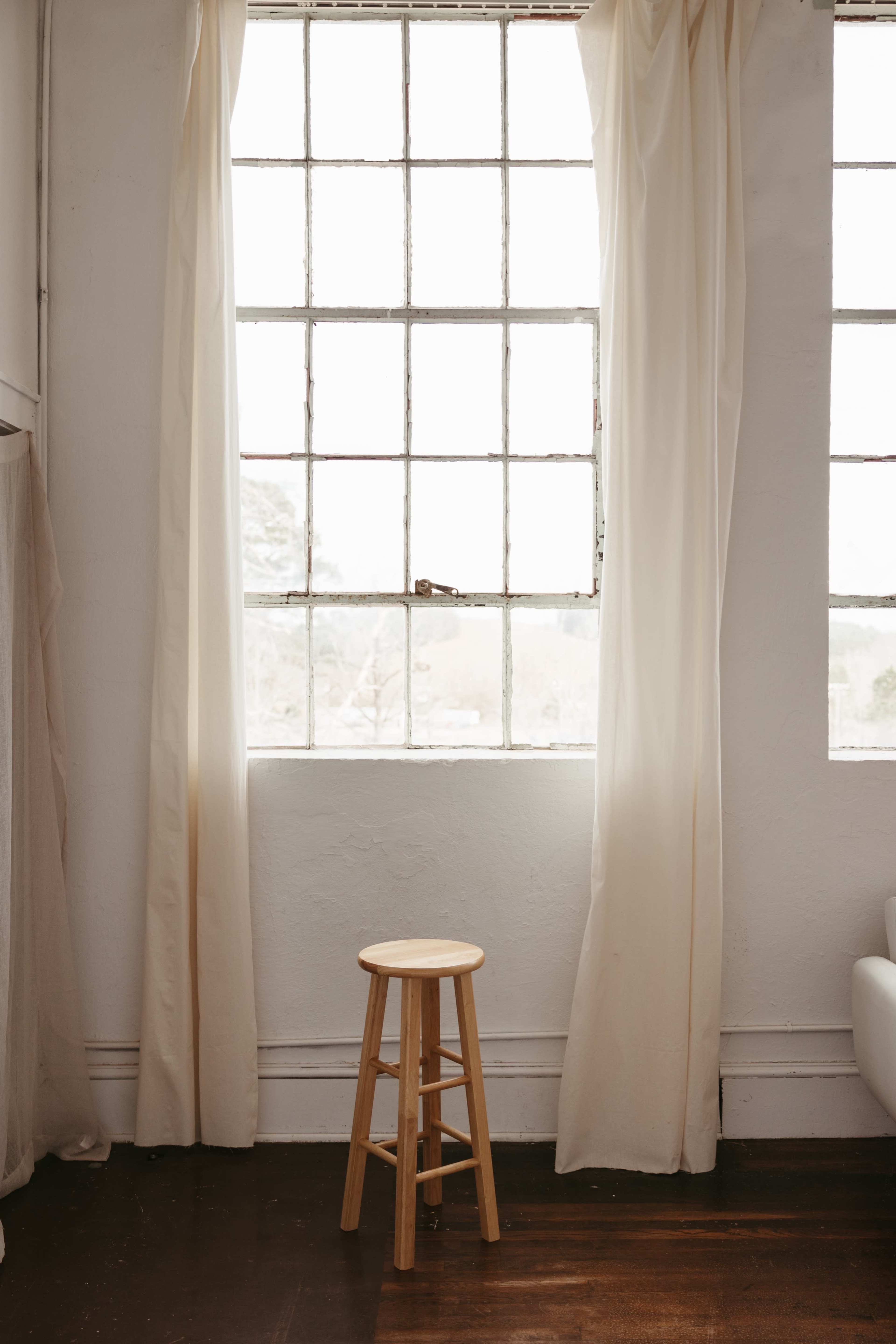A wooden stool sits in front of a large window with light fabric curtains, allowing soft light to fill the room.