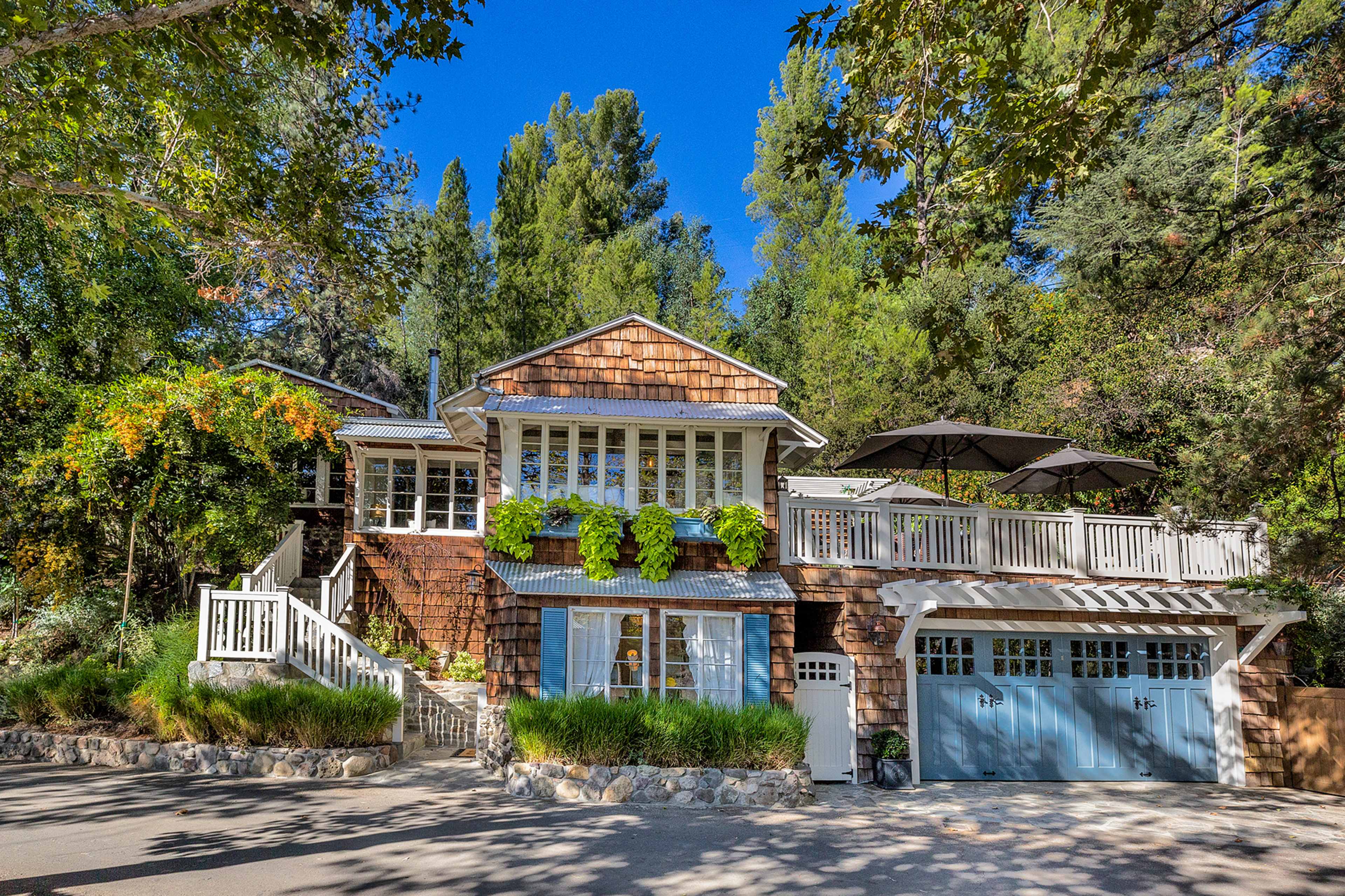 A two-story house with a wooden exterior and a balcony surrounded by greenery sits in a wooded area.