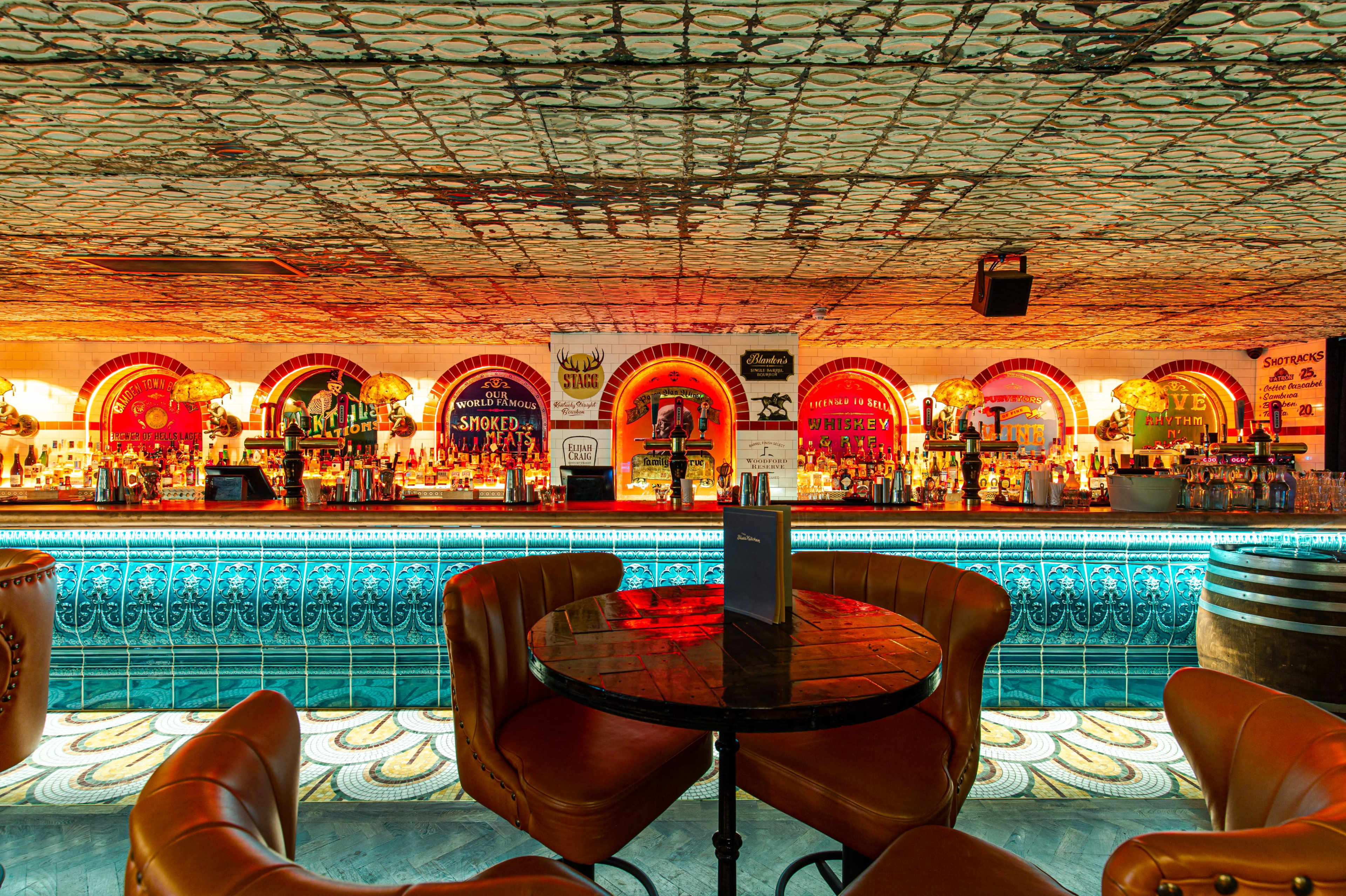 The image shows a bar interior with illuminated shelves of liquor, circular booths, and a patterned tiled floor.