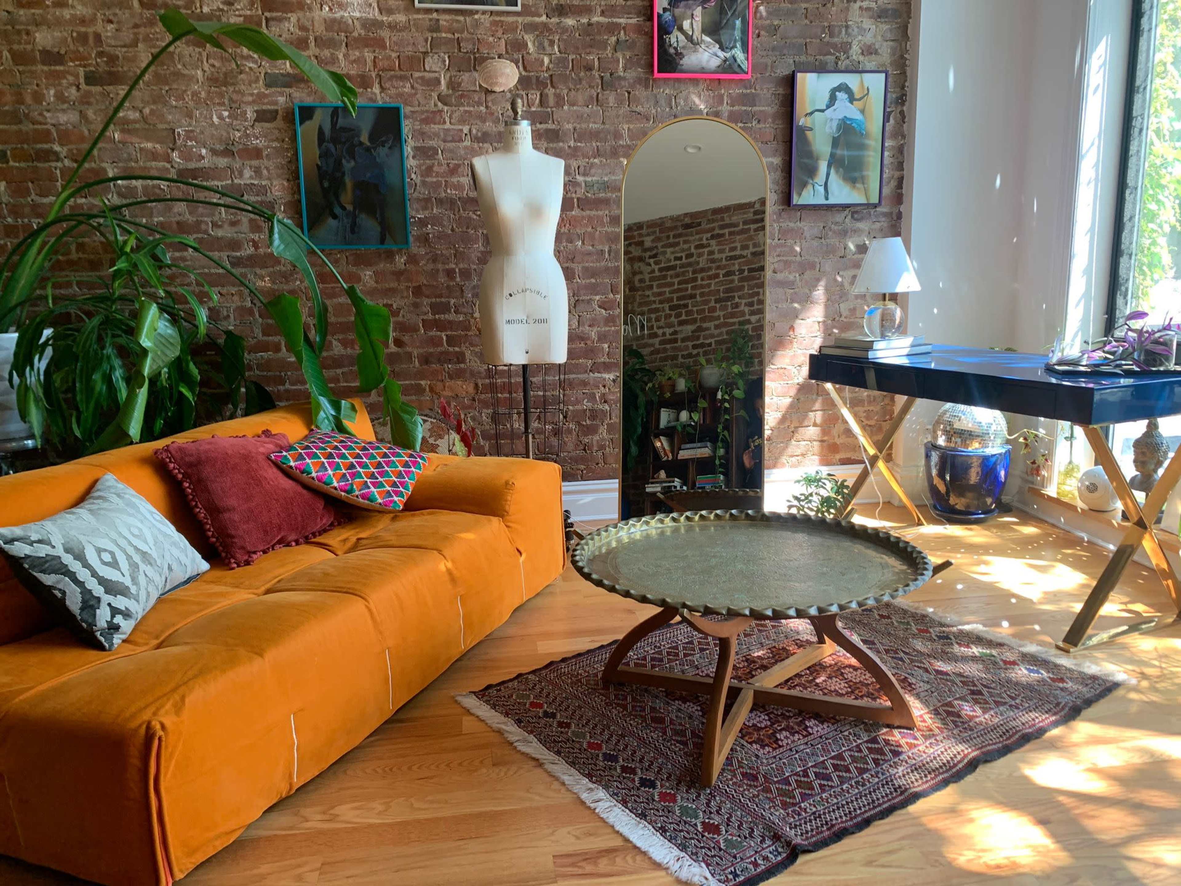The image shows a cozy room featuring an orange sofa with decorative pillows, a round coffee table on a patterned rug, a mannequin, and various wall art against a brick backdrop.