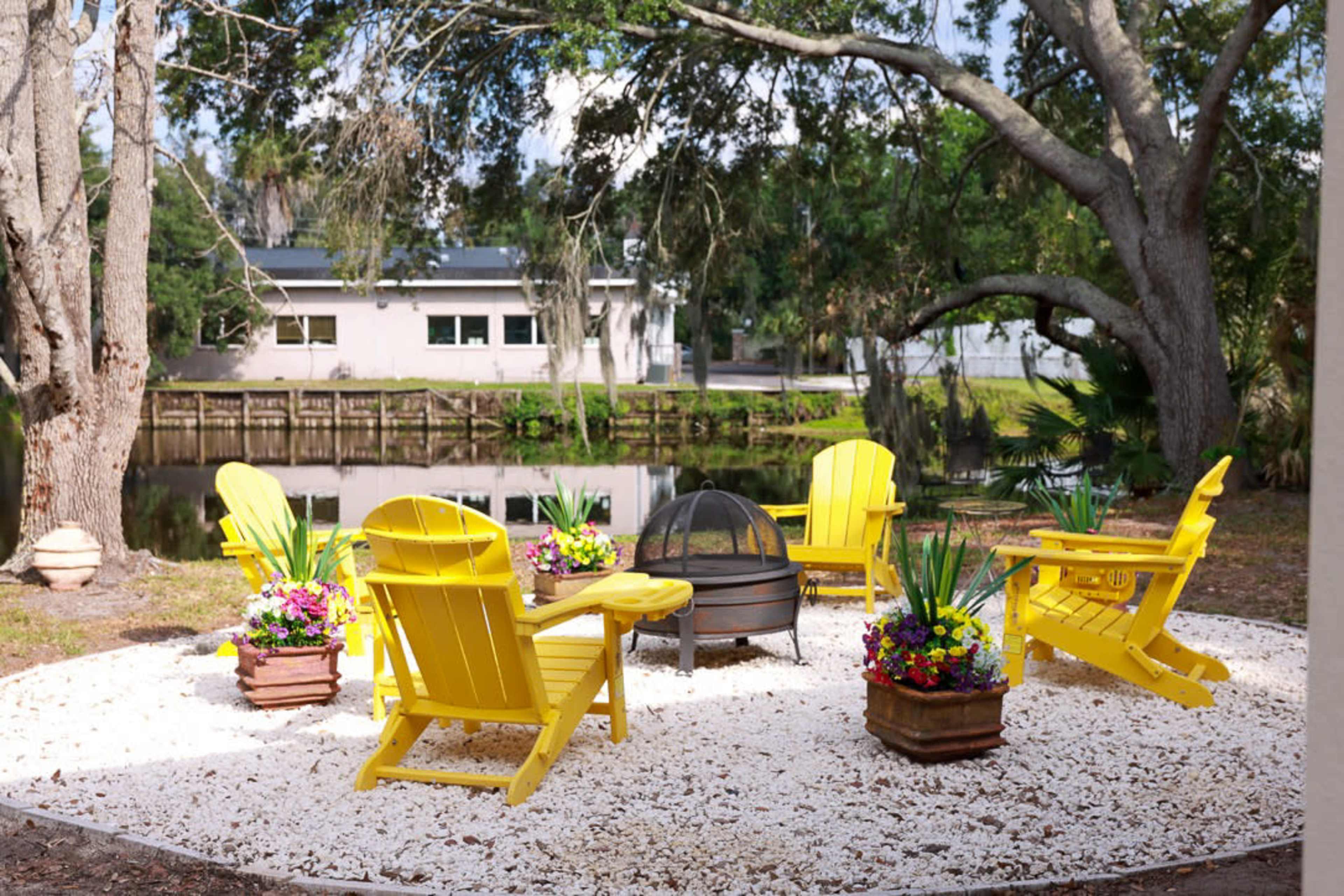 The image shows a circular seating area with six yellow Adirondack chairs arranged around a fire pit, surrounded by flower pots and located near a body of water.
