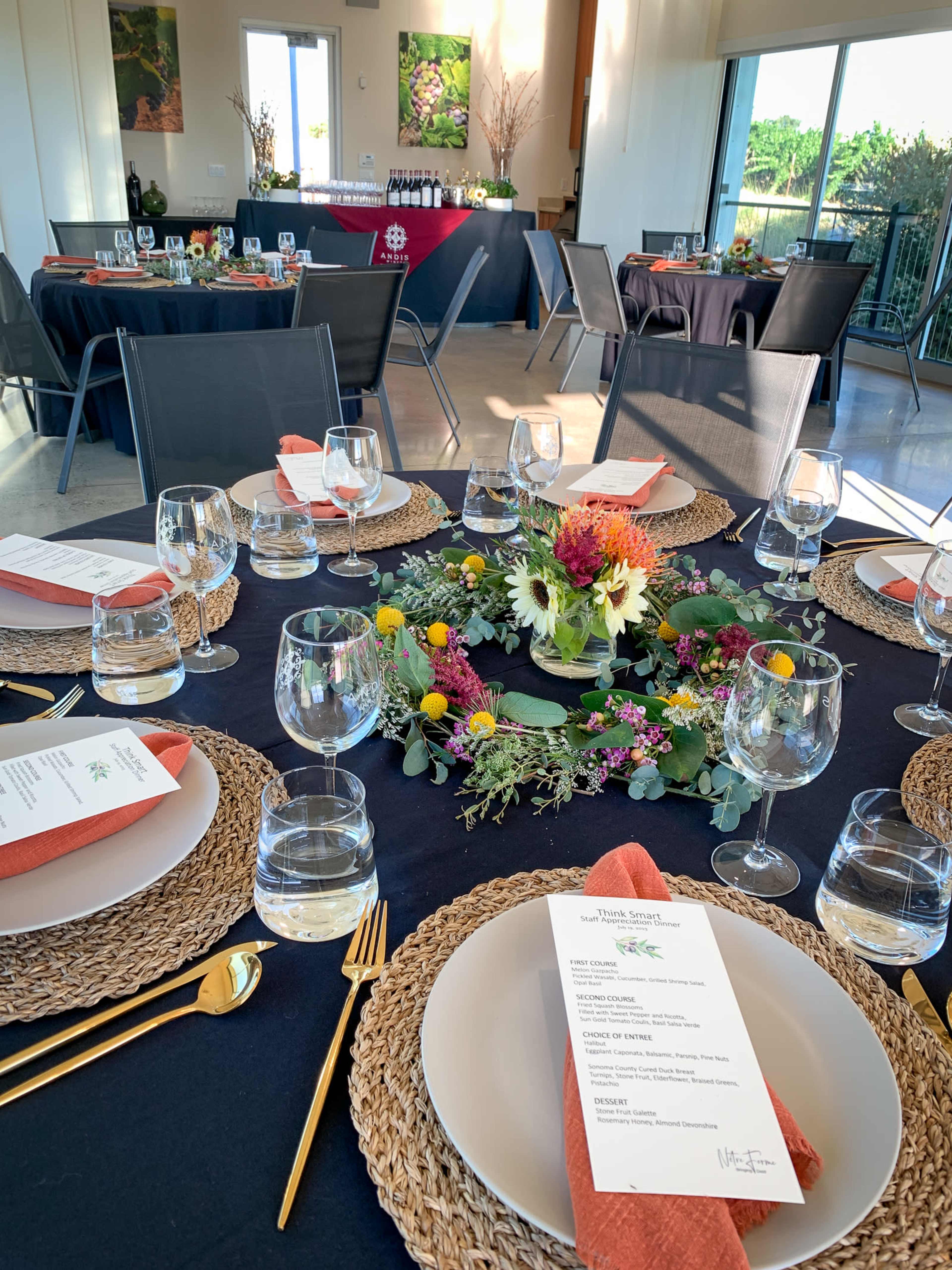 A dining setup is arranged with tables featuring woven placemats, glassware, and floral centerpieces, set in a bright room that opens to an outdoor view.
