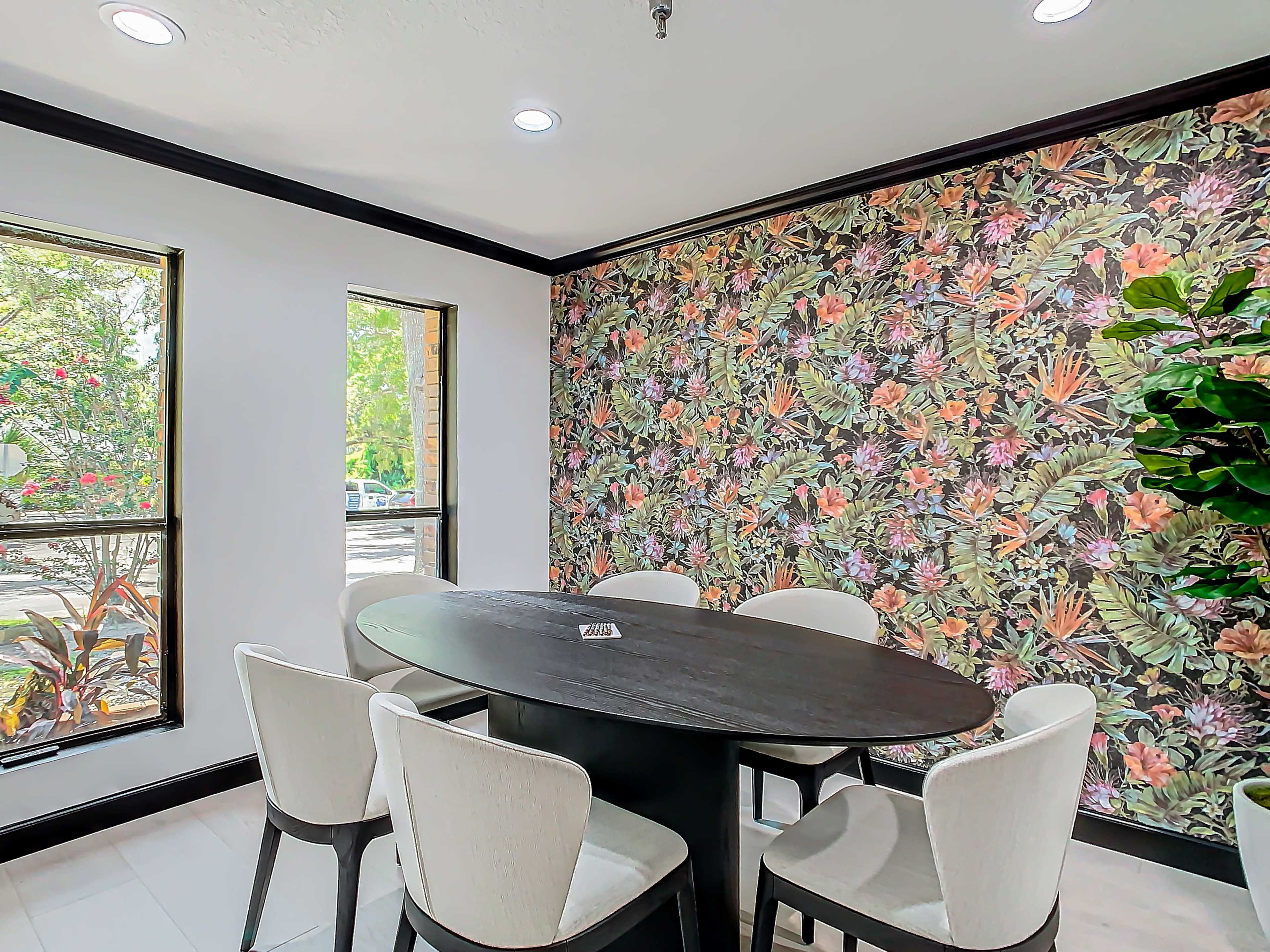 A dining area features a round black table surrounded by white chairs, with floral wallpaper and windows letting in natural light.