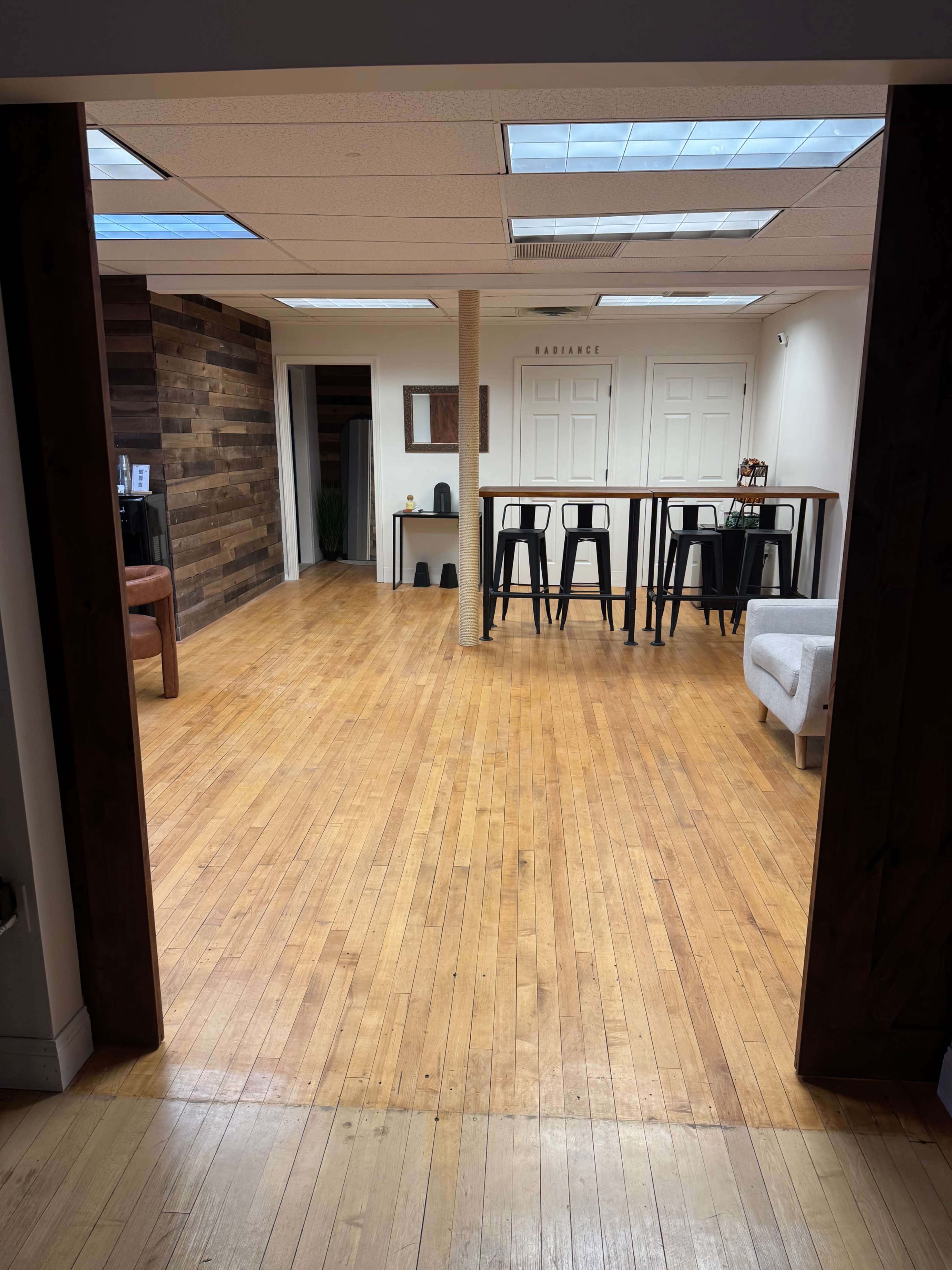 The image shows a spacious, well-lit office with wooden floors, a central column, and furniture such as bar stools and a sofa.