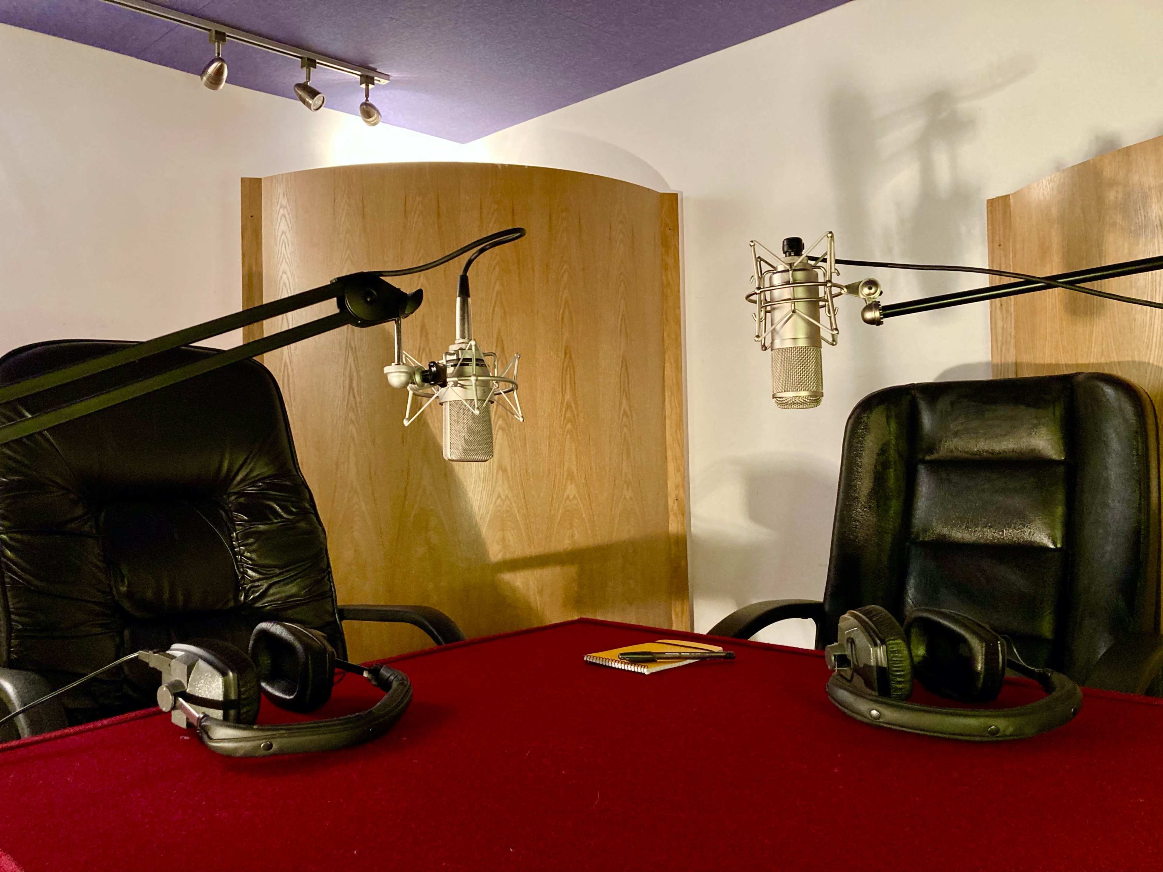 A recording studio setup with two microphone stands, two black chairs, and headphones resting on a red table.