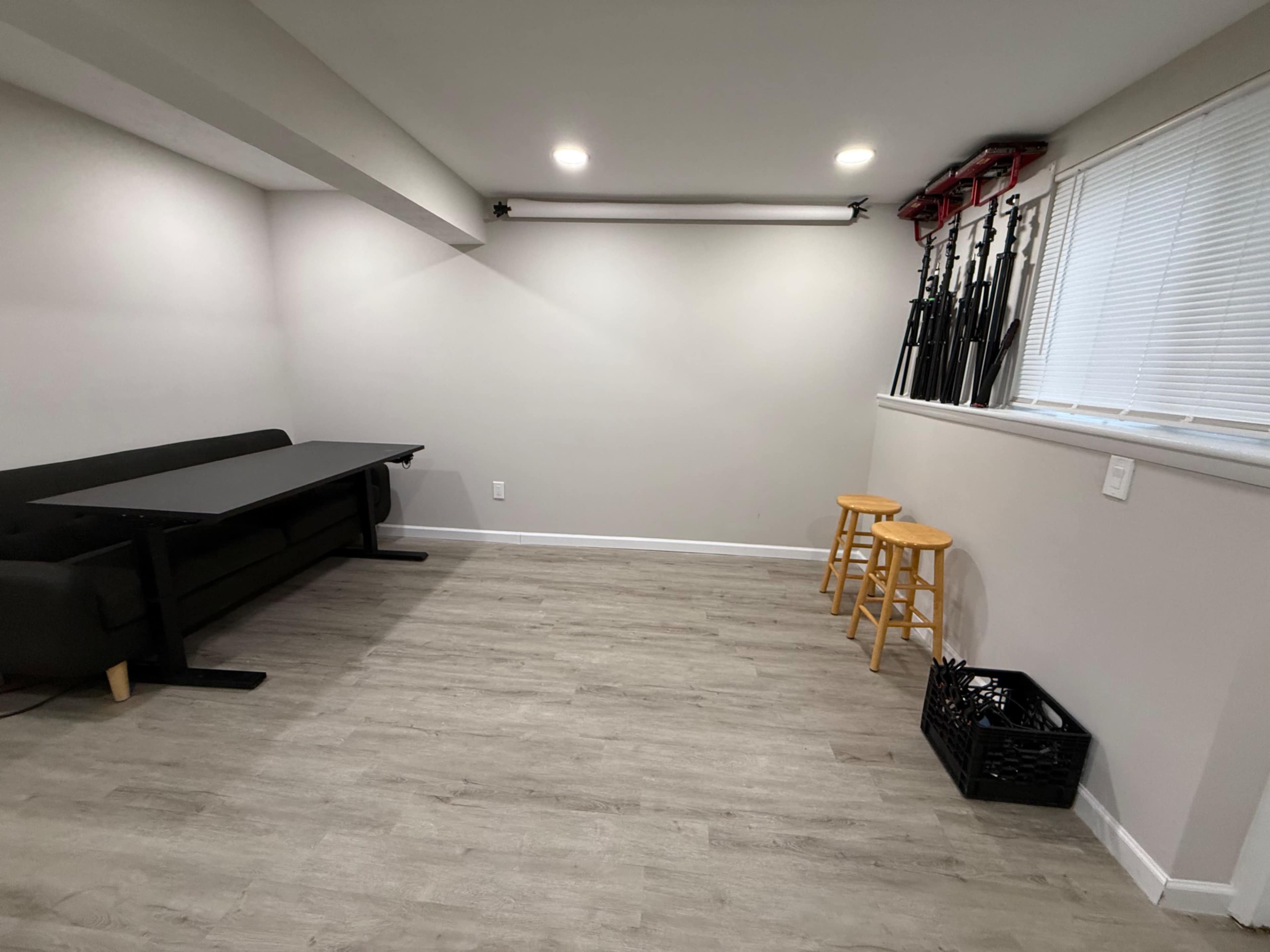 The image shows a sparsely furnished room with a black couch, a folding table, two wooden stools, and various tripods mounted on the wall.