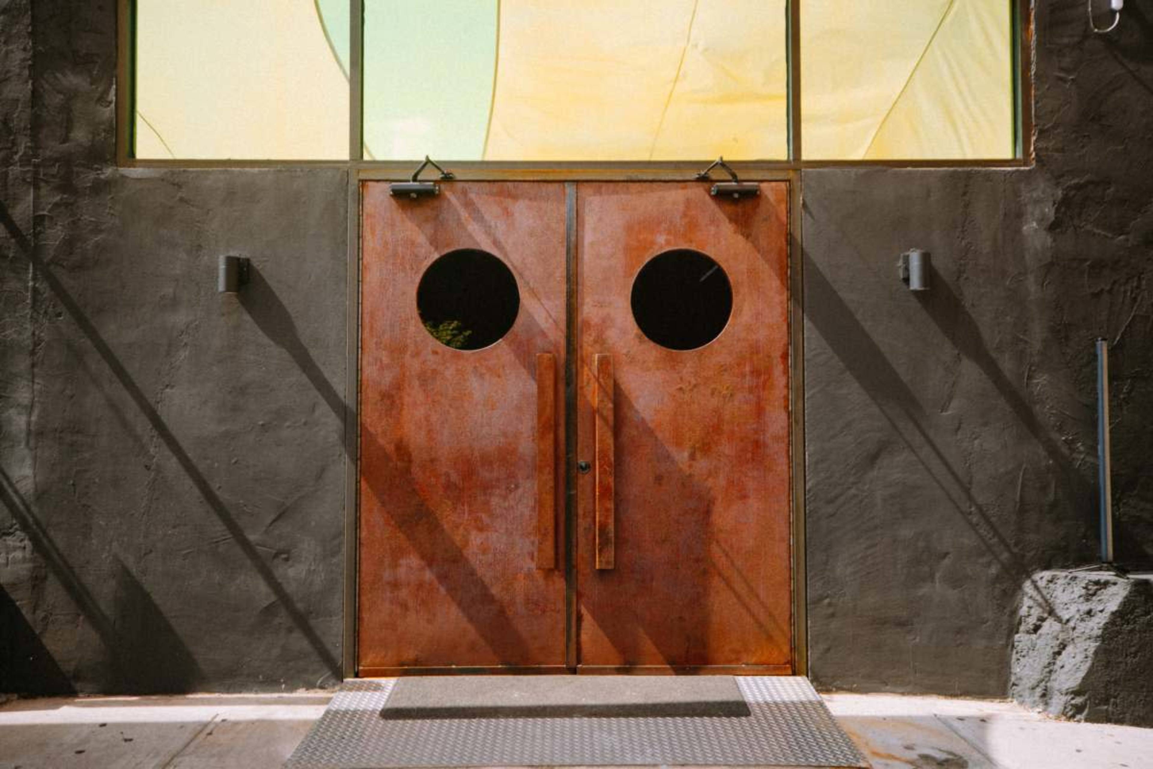 A pair of circular-doored metal entrance doors is installed in a textured gray wall, with light filtering through a large window above.