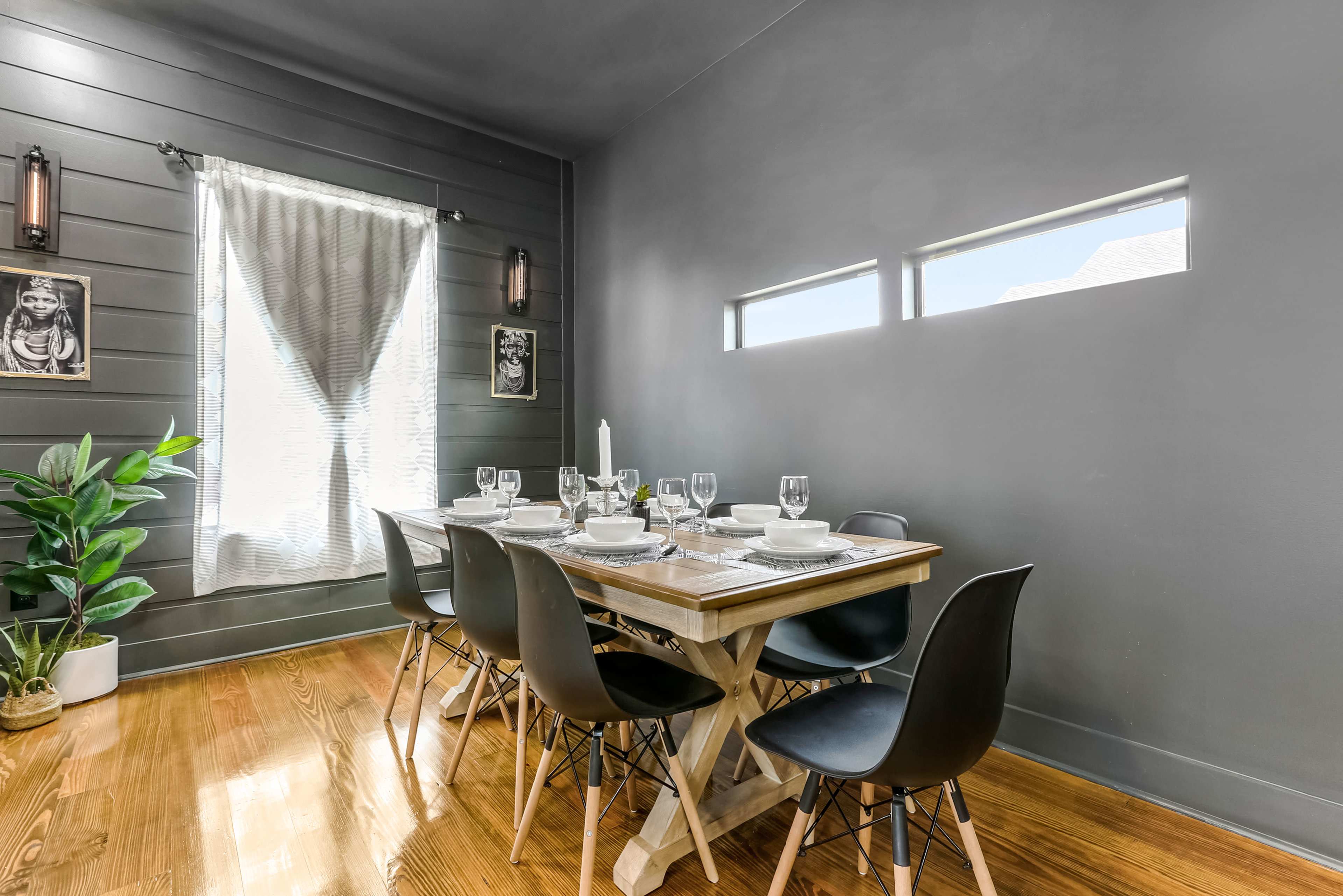 A dining room features a wooden table set for six, surrounded by black chairs, with a large window letting in natural light and decorated walls.