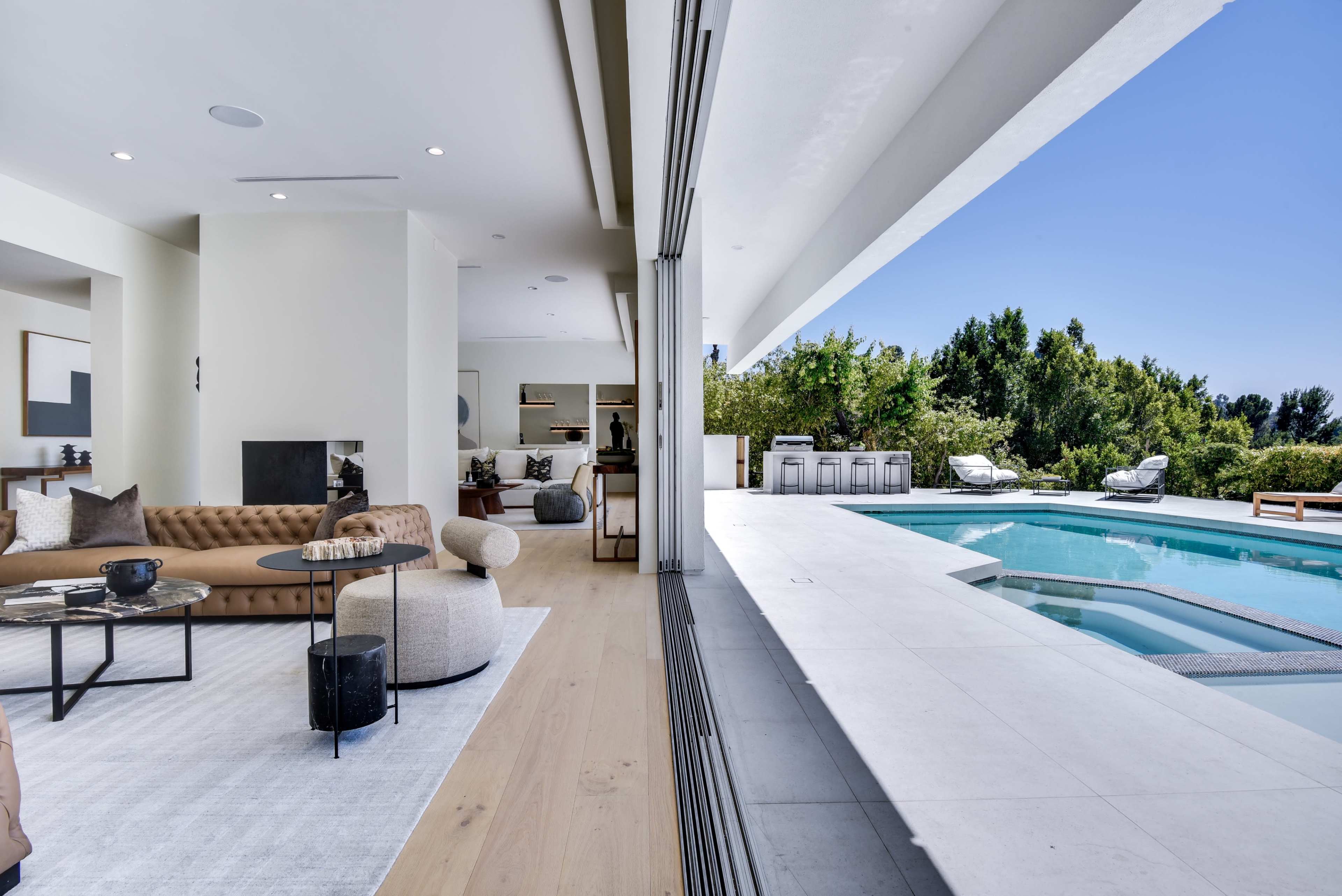 A modern living room opens to a sleek outdoor pool area surrounded by lush greenery.