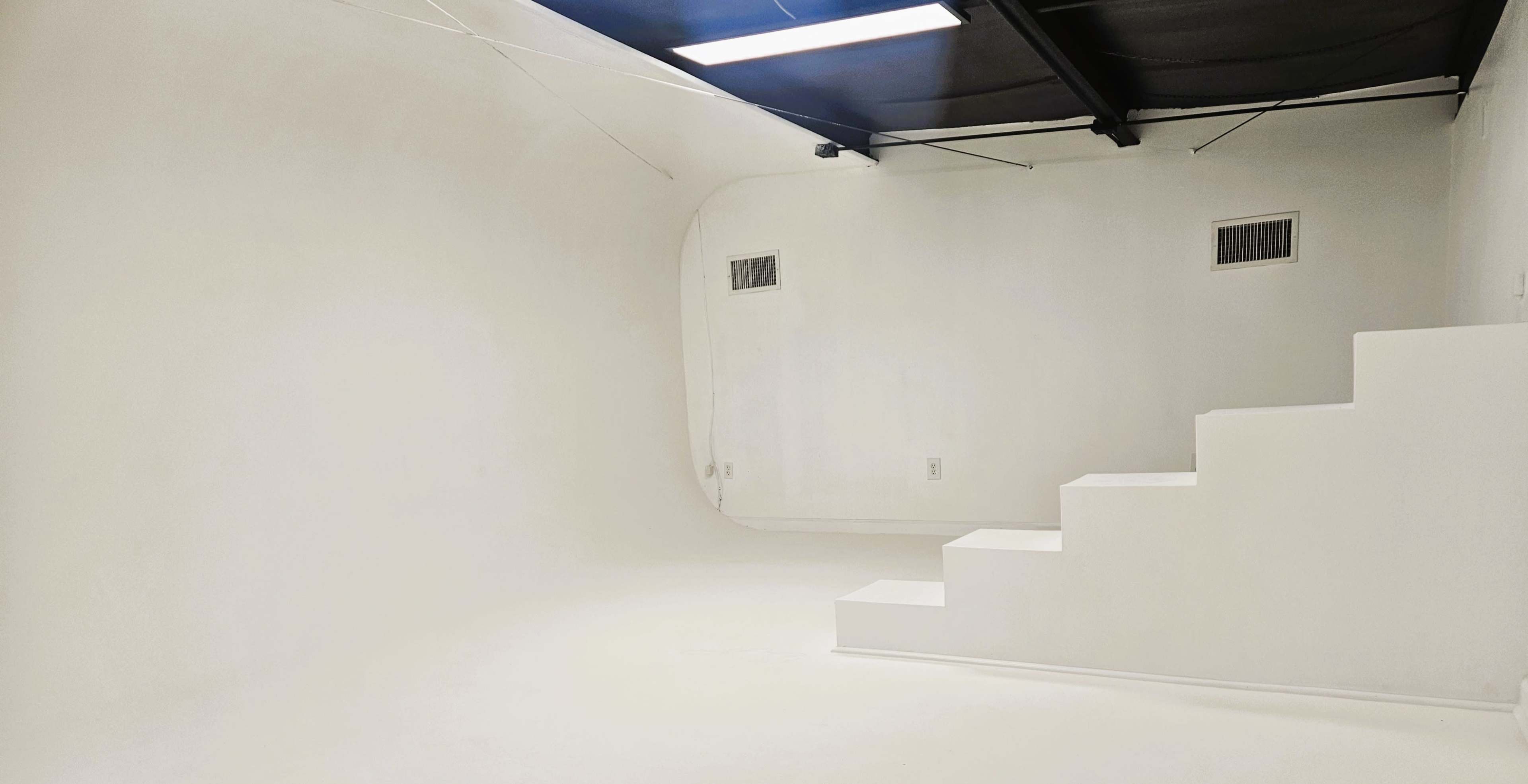The image shows a minimalistic white room featuring a curved wall and a staircase leading to a small elevated platform.