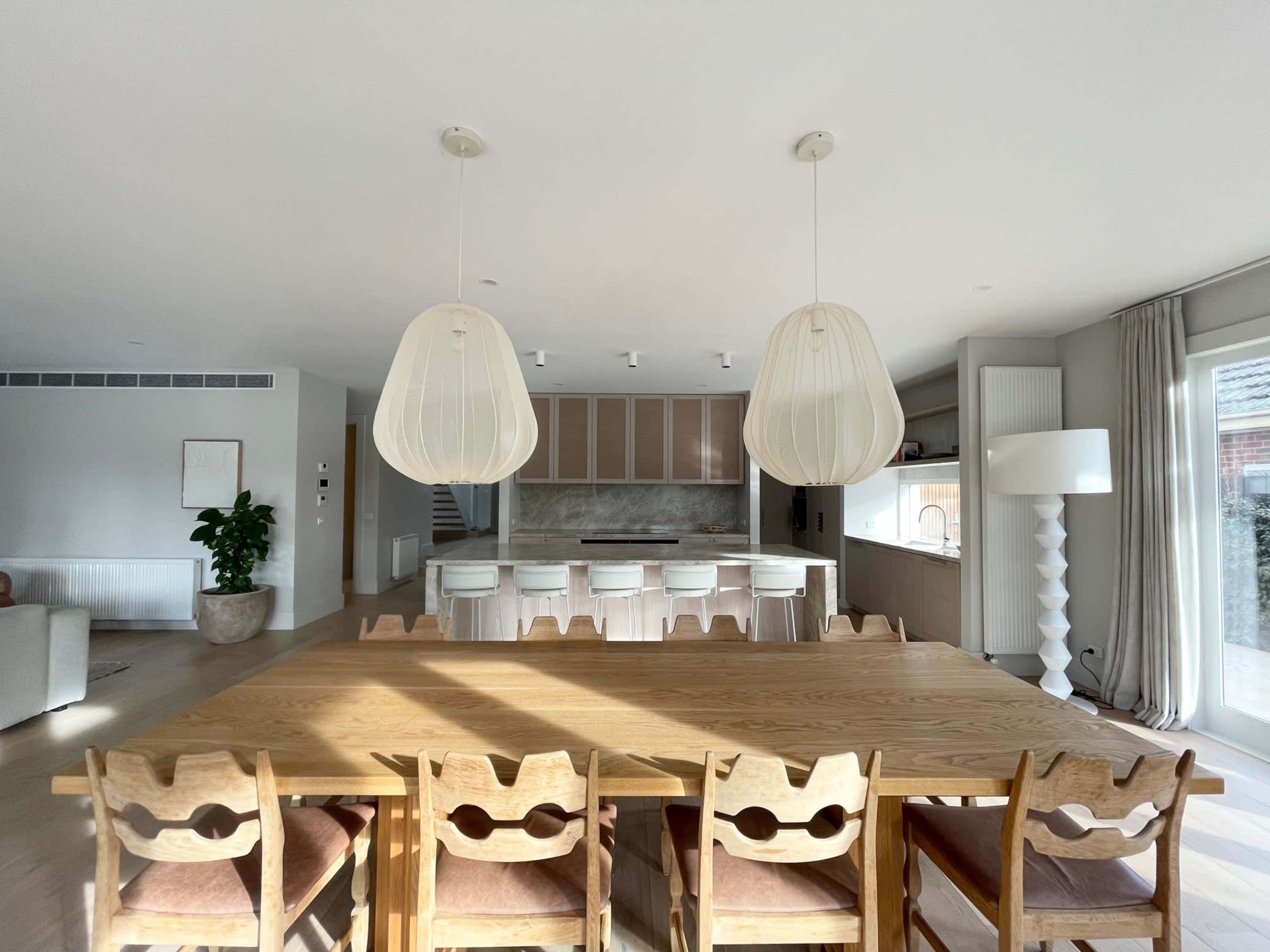 A spacious, modern kitchen and dining area features a long wooden table with eight chairs, large pendant lights overhead, and a minimalist design.