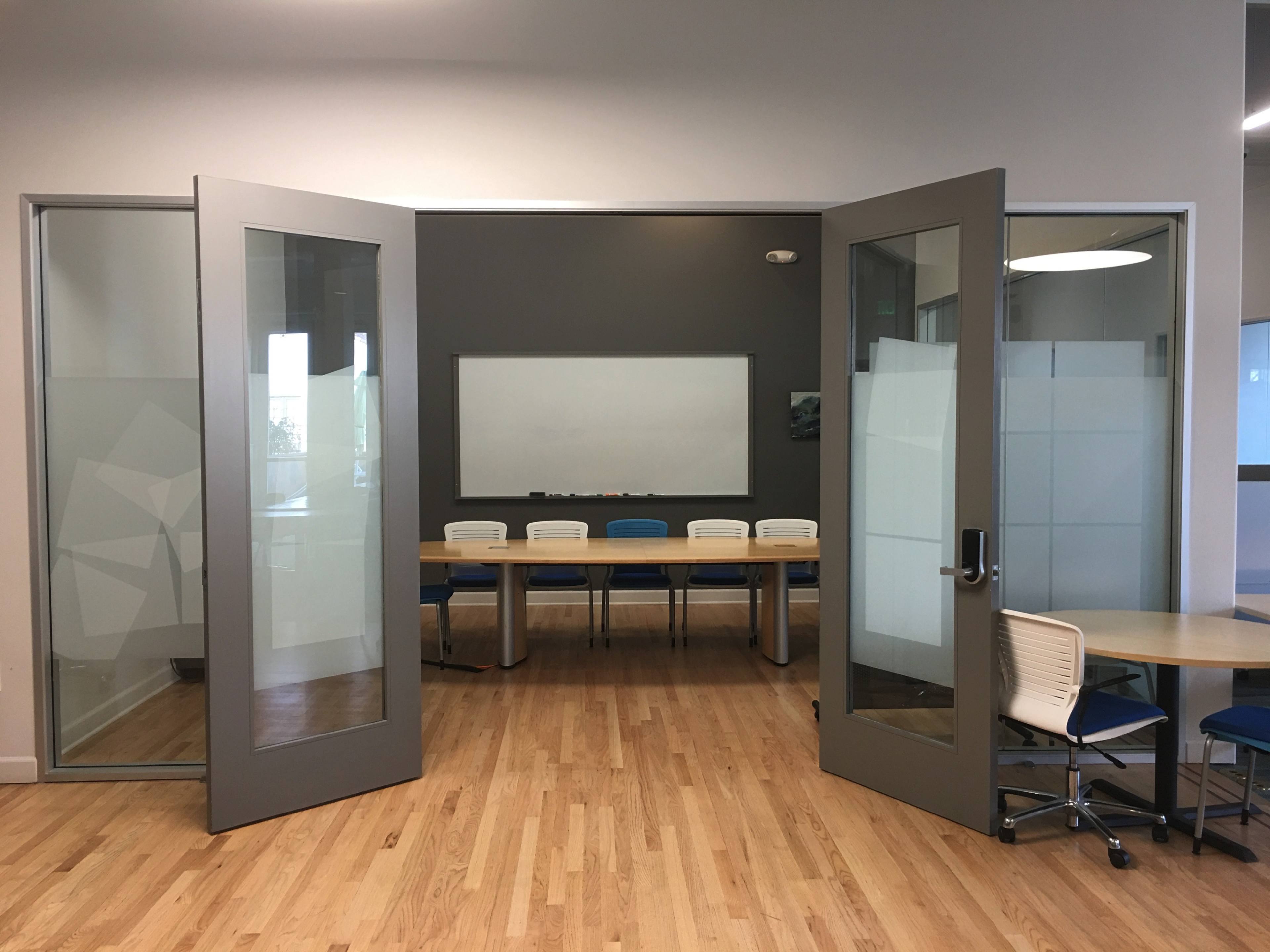 The image shows a conference room with a large wooden table and white chairs, accessible through double glass doors.