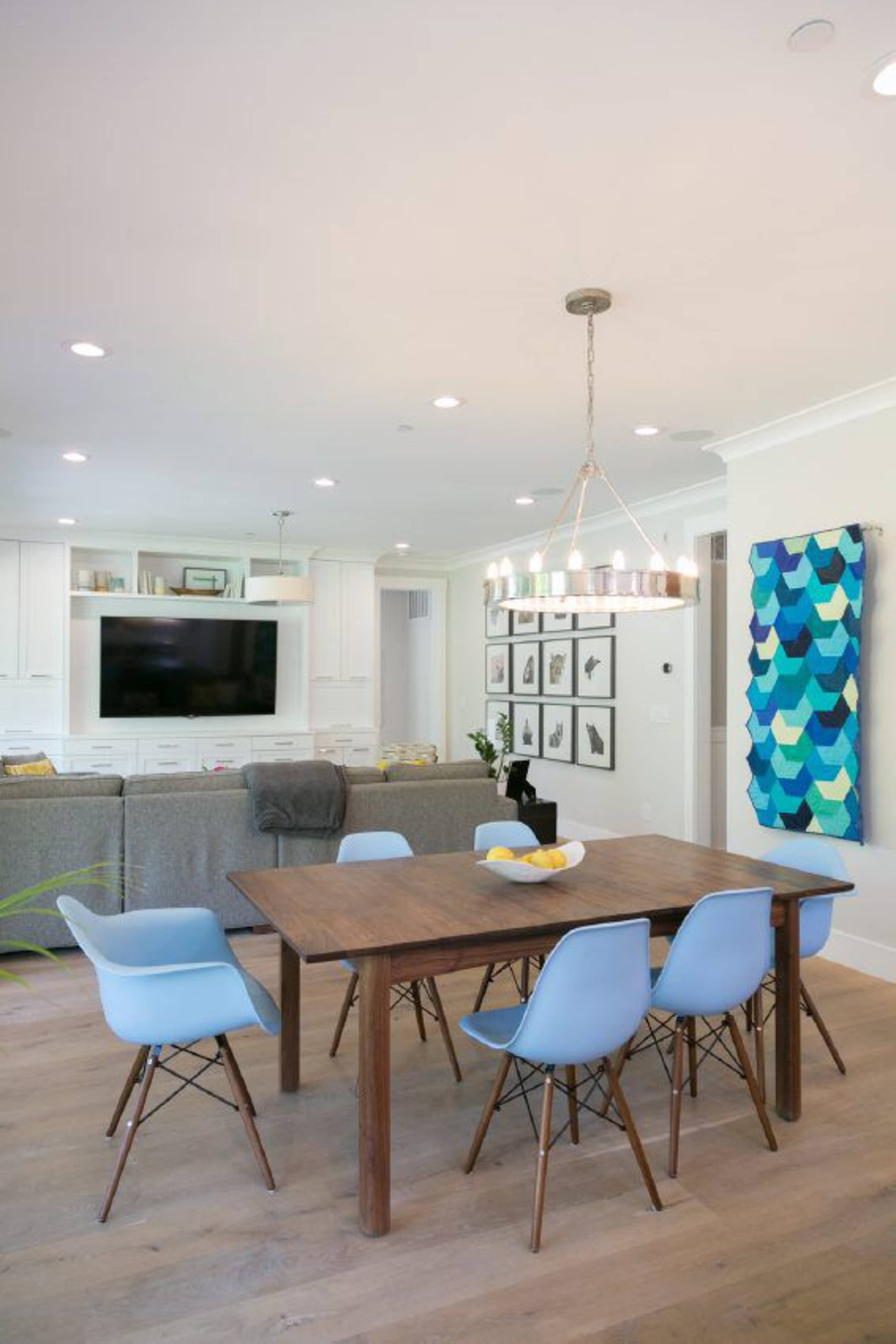 A modern dining area featuring a wooden table surrounded by five blue chairs, with a television, artwork, and a sofa in the background.