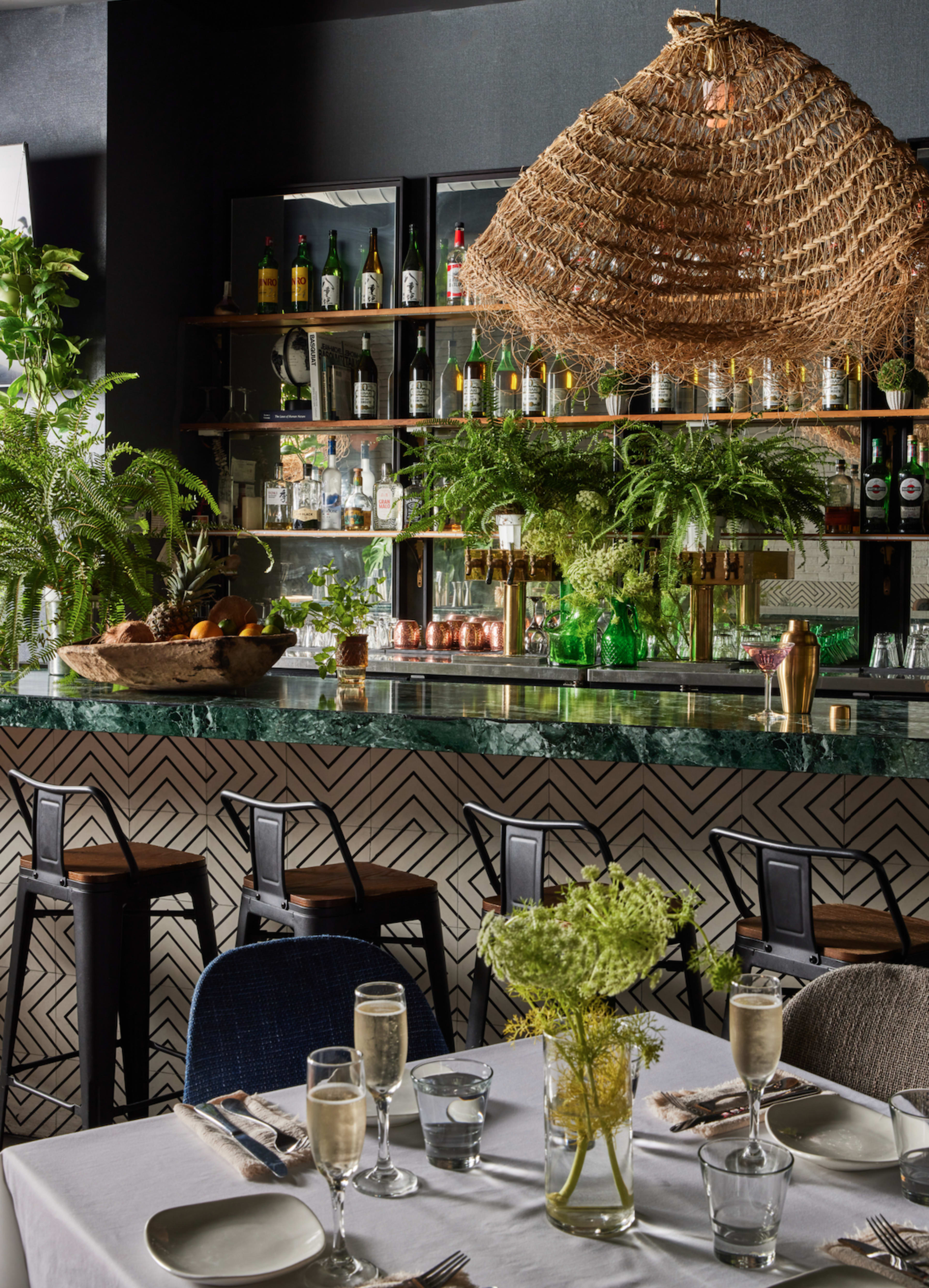 The image shows a stylish bar area in a restaurant, featuring a woven pendant light, a polished counter with green accents, and shelves lined with bottles and greenery.