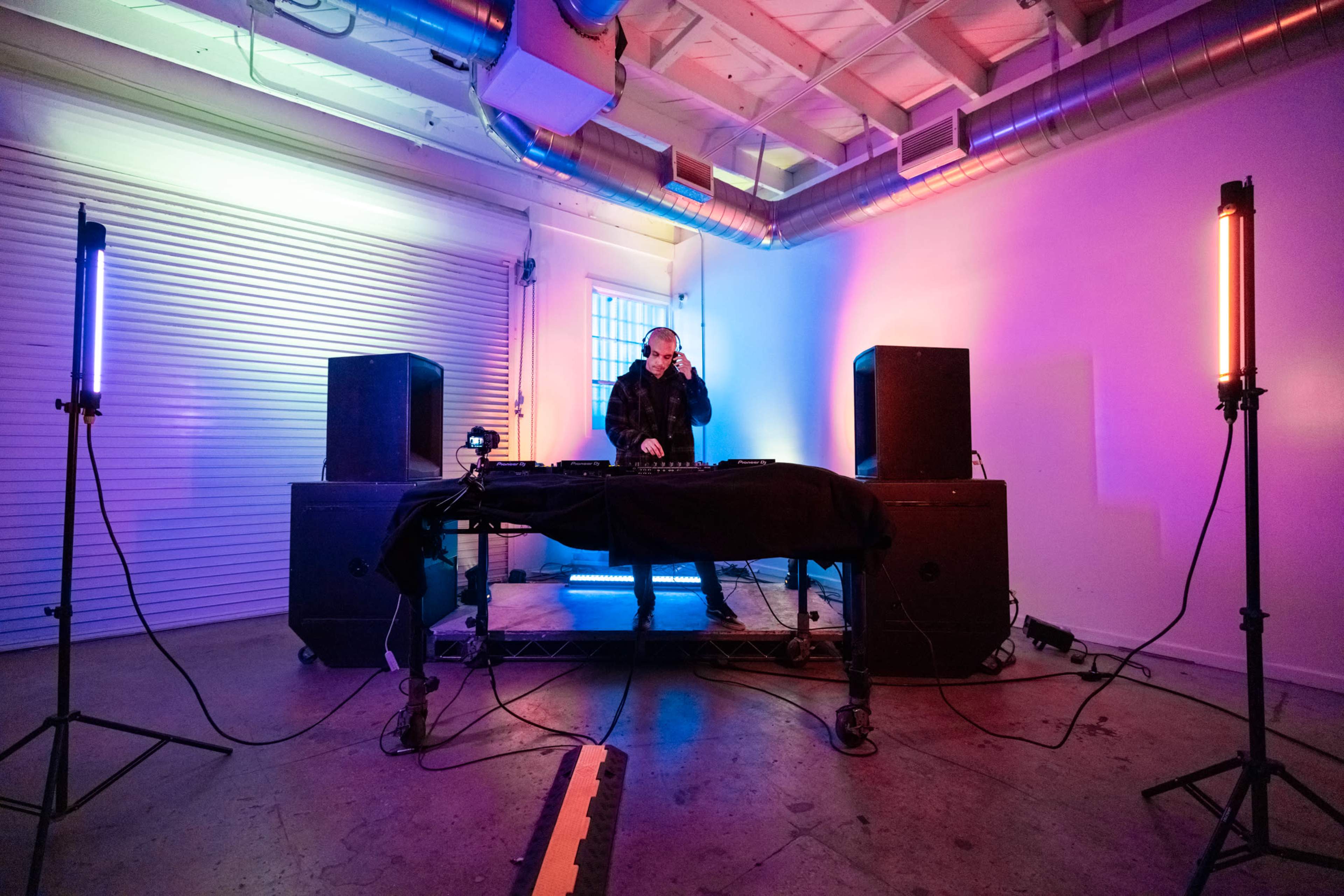 A DJ performs at a setup with colorful lighting in a sparse indoor space.