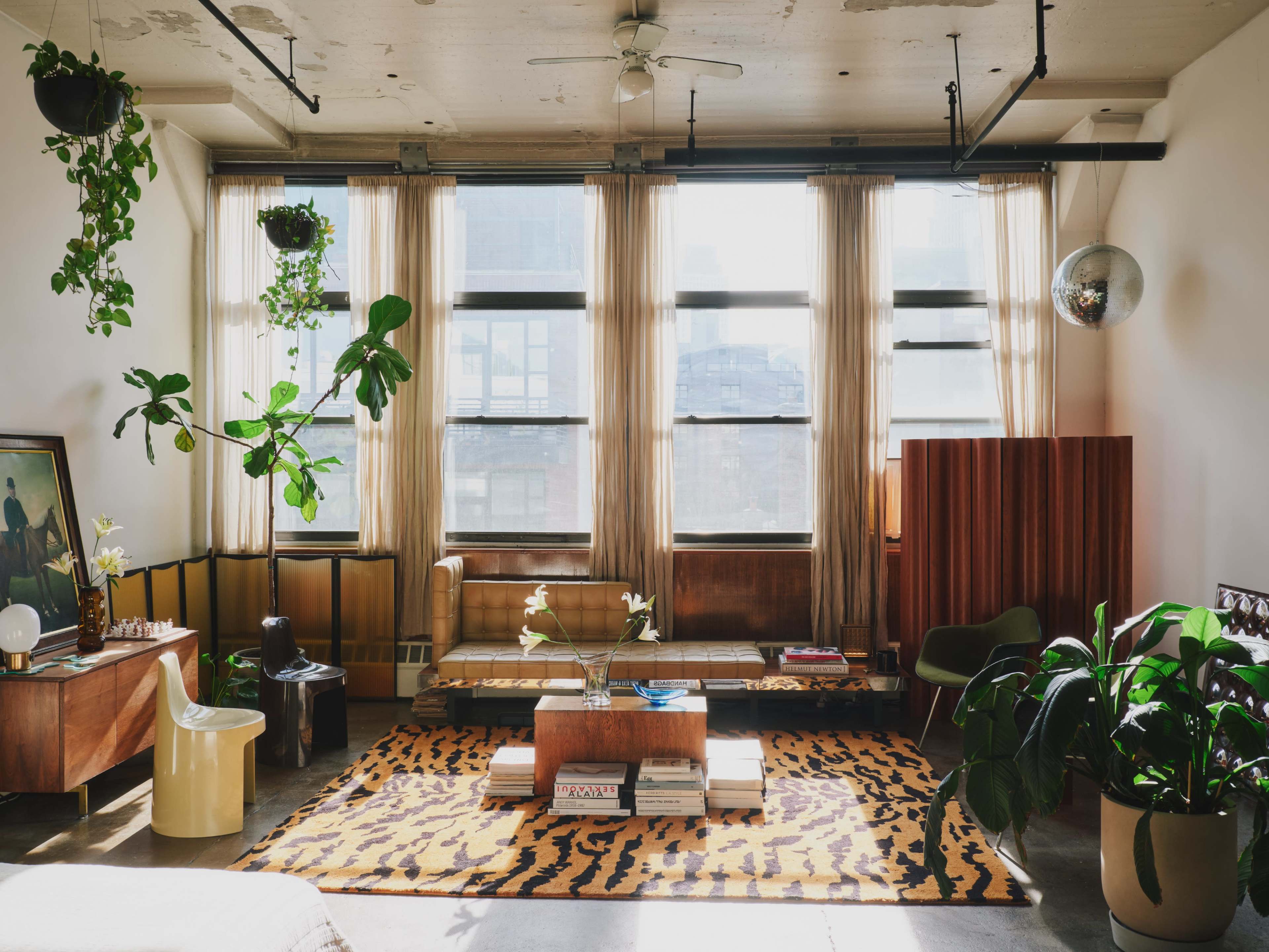 The image depicts a bright, spacious living room with large windows, plants hanging from the ceiling, and a mix of modern and vintage furniture arranged around a patterned rug.