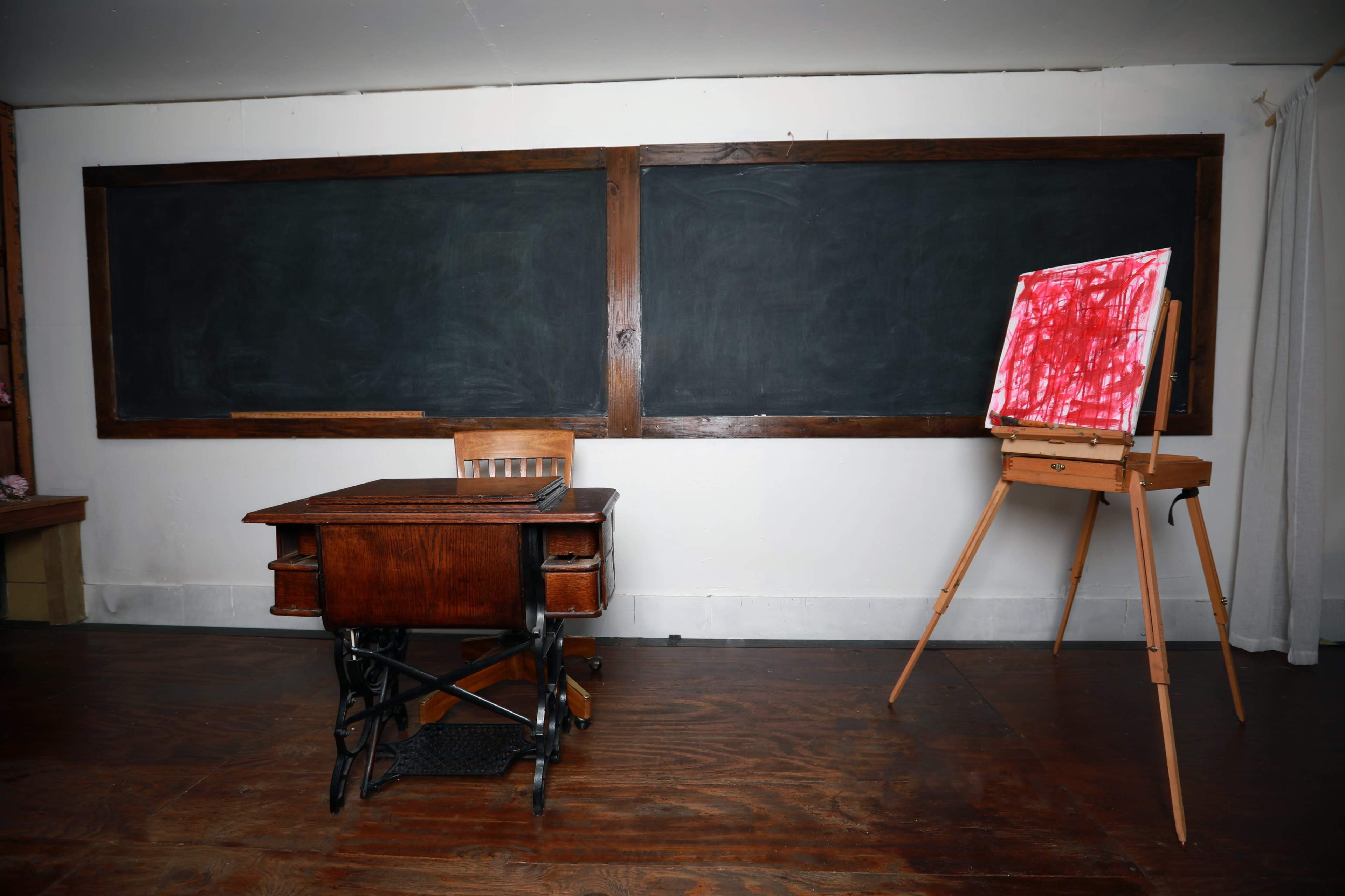 A wooden classroom desk in front of two chalkboards, with an easel displaying a canvas covered in red paint on the right.
