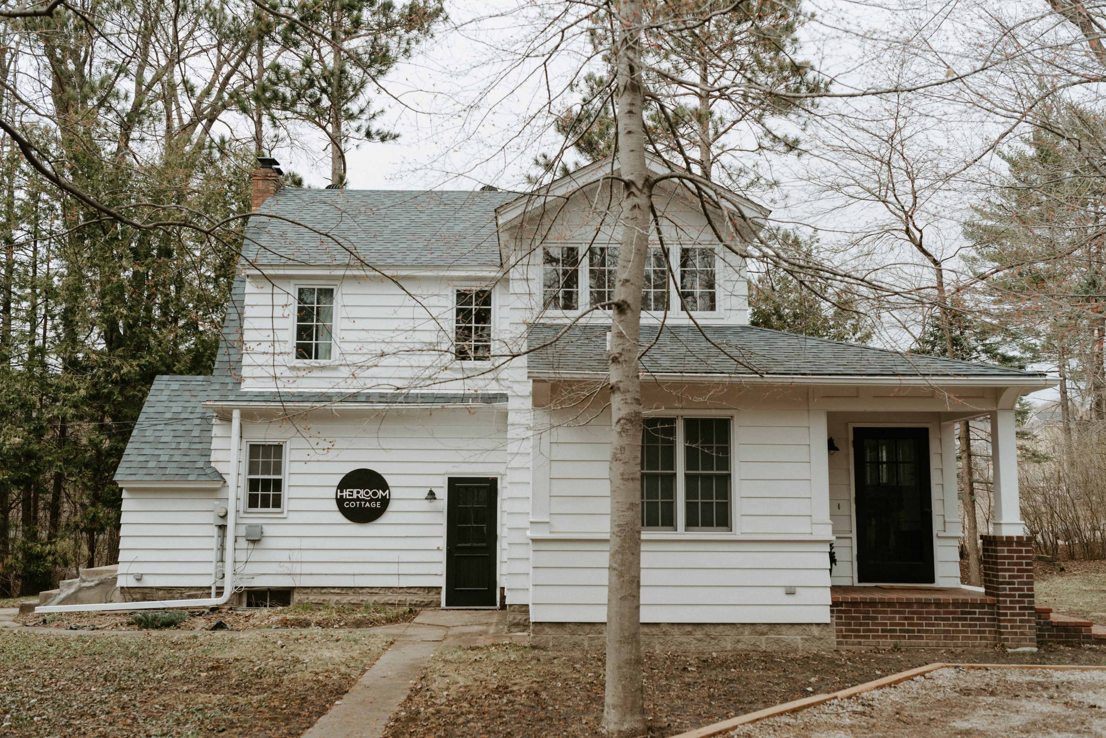A two-story white house with a steeply pitched roof and dark green doors sits in a treed yard.