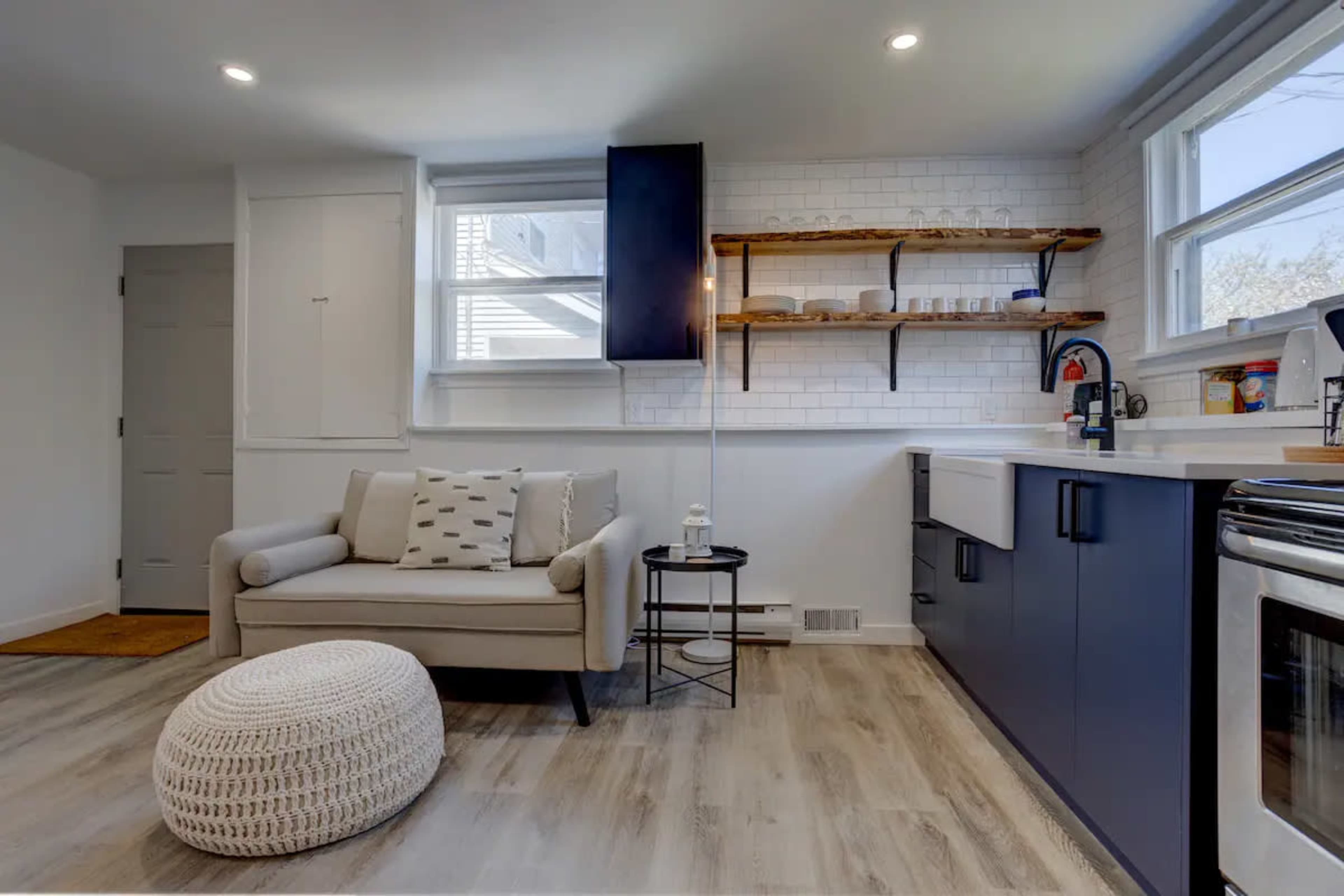 A modern kitchen with white walls, a blue cabinetry unit, open shelving, a small couch, and a round pouf.