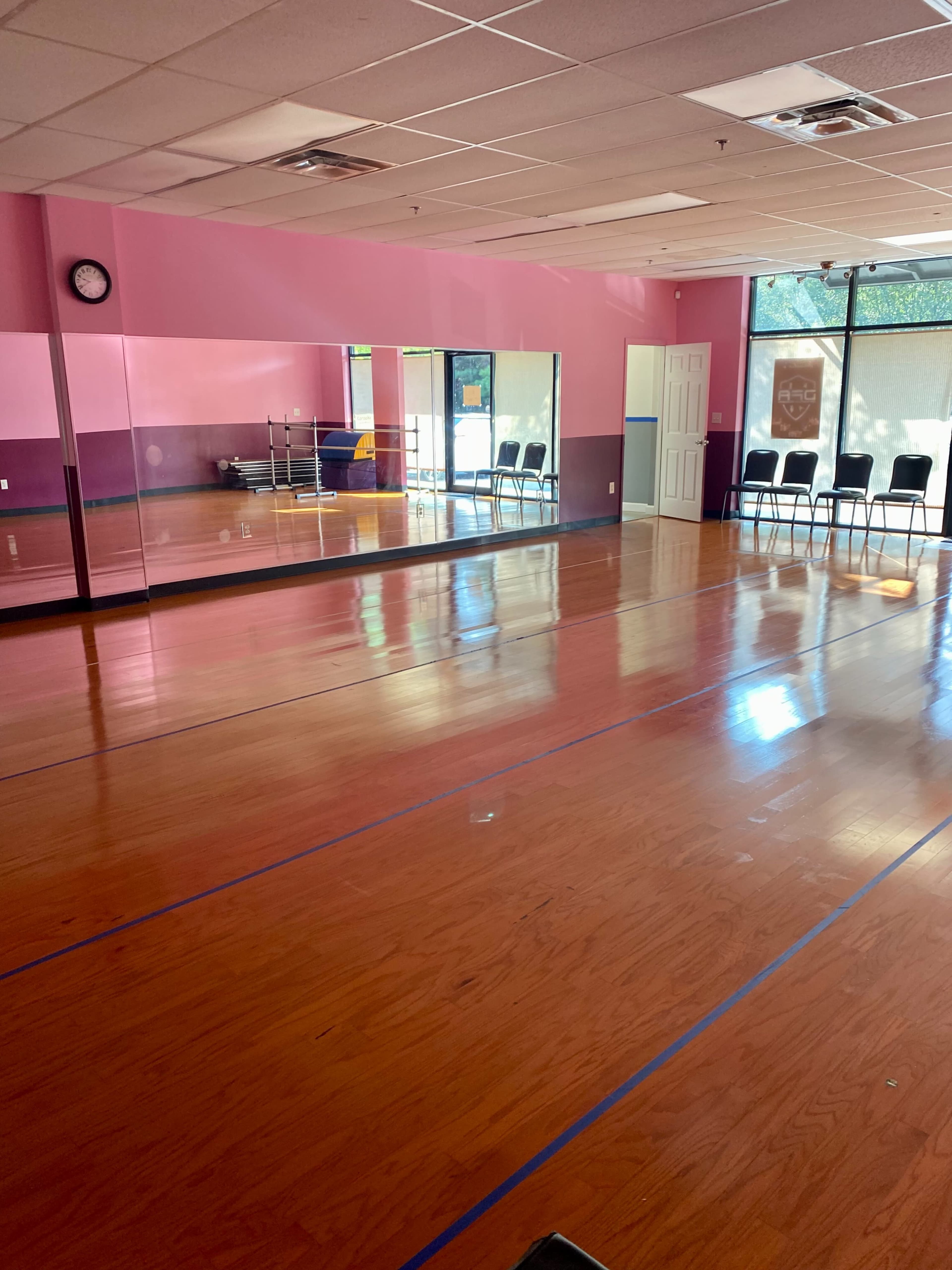 A spacious dance studio with hardwood floors, pink walls, large mirrors, and a set of chairs lined against the wall.