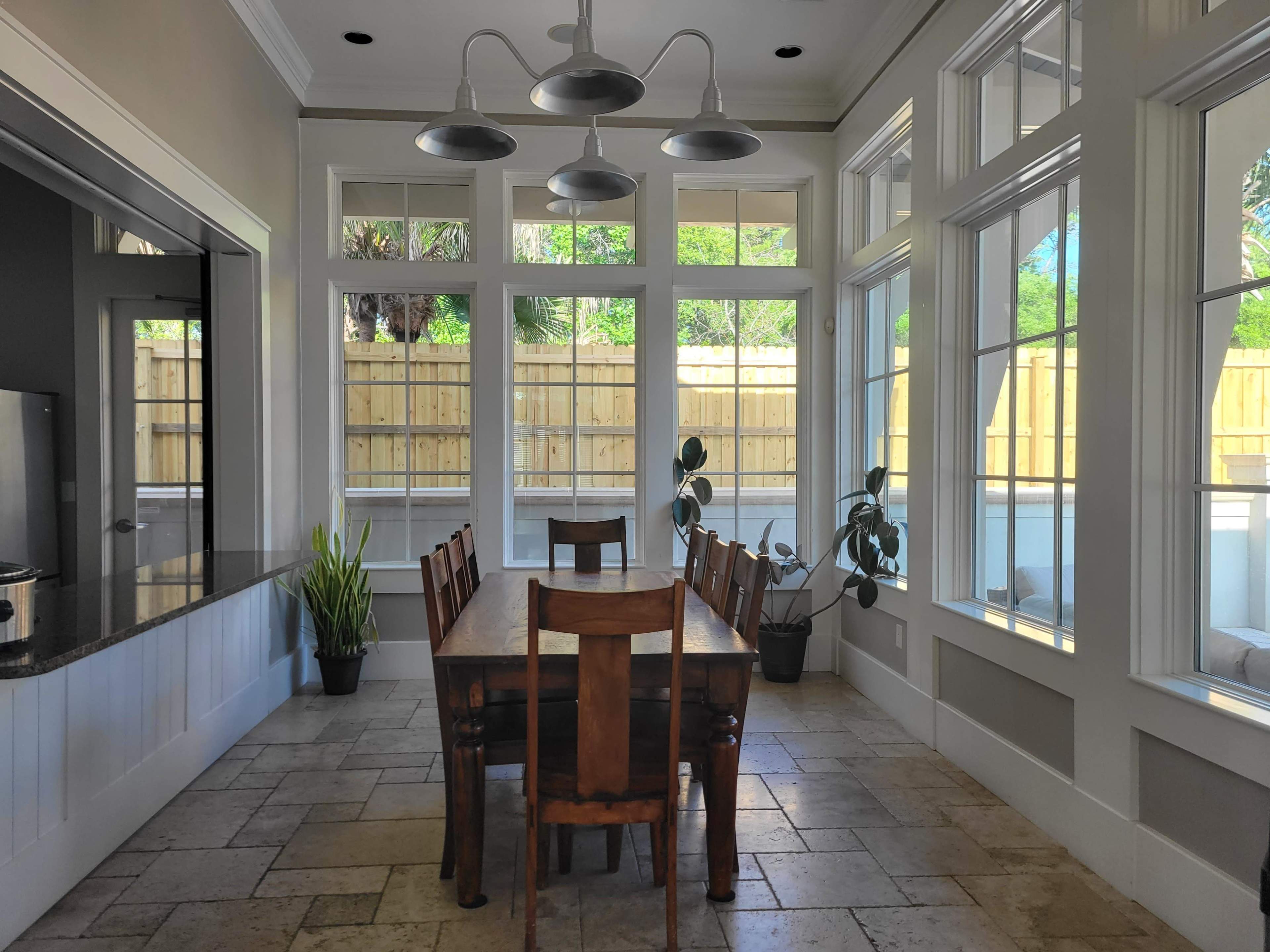 A well-lit dining area features a wooden table surrounded by chairs, with large windows and a view of a fenced outdoor space.