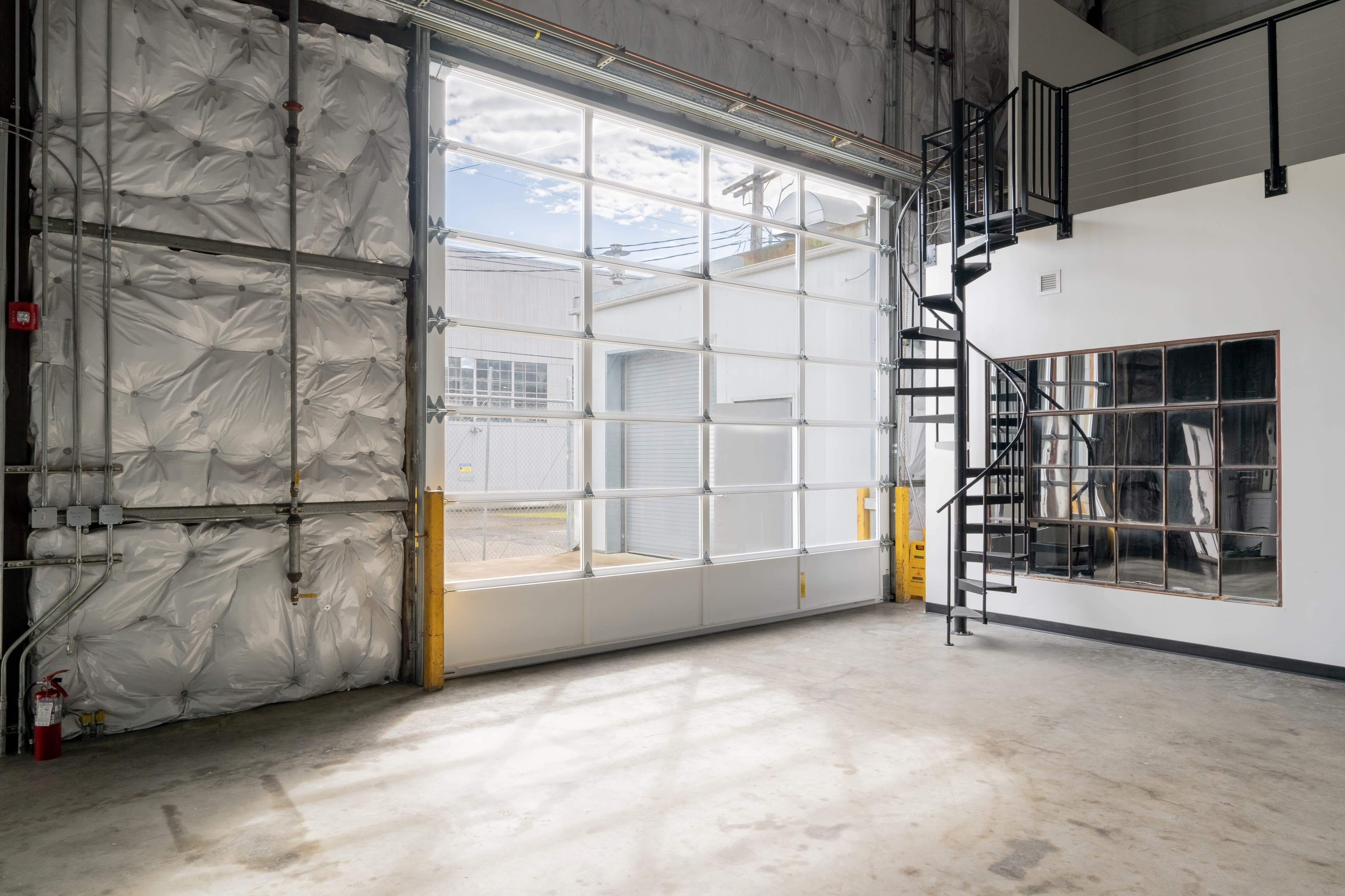 The image shows an industrial space featuring a large glass garage door, a metal spiral staircase, and exposed walls with insulation.