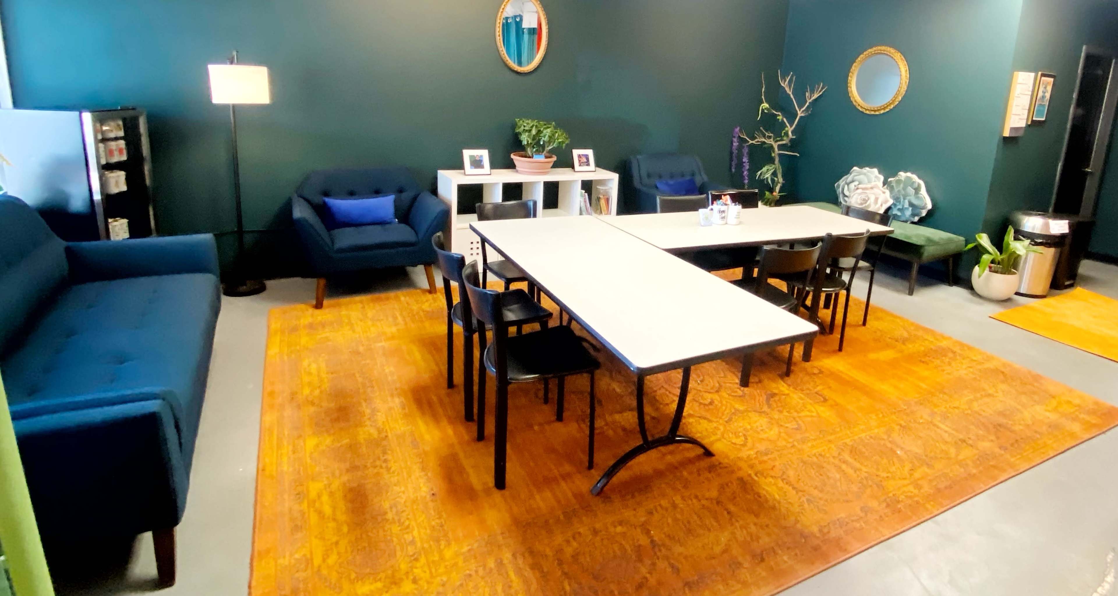A meeting room with a long table surrounded by chairs, two sofas, decorative plants, and mirrors on the dark green walls.