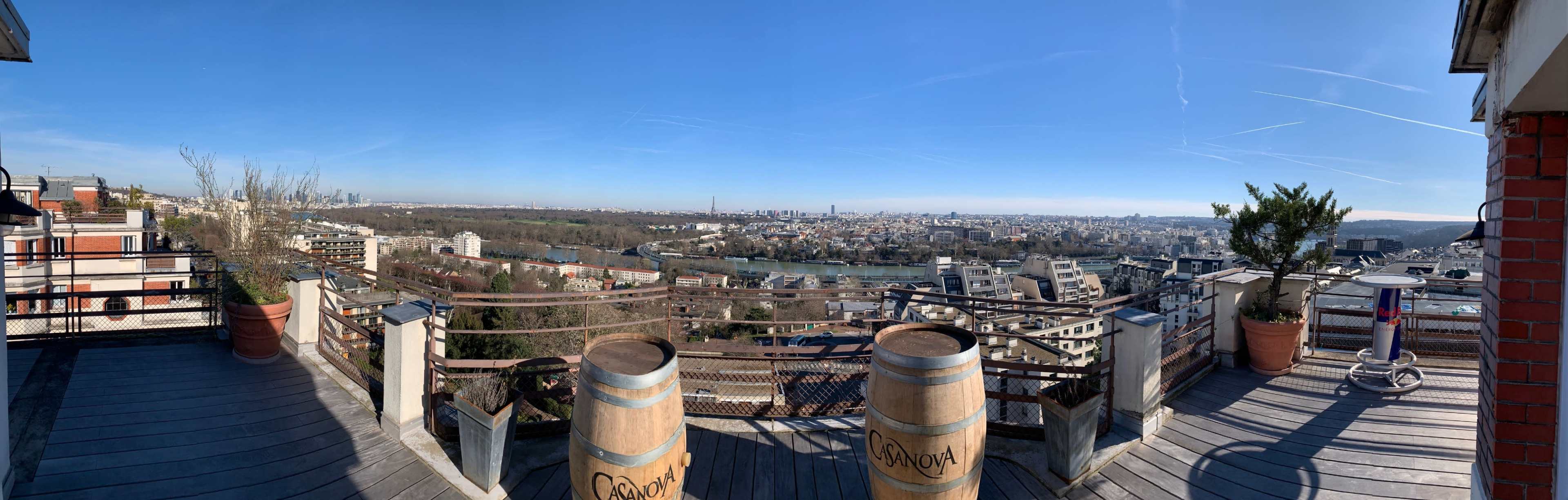 A panoramic view of a city skyline next to a river, taken from a rooftop terrace lined with wooden barrels and planters.