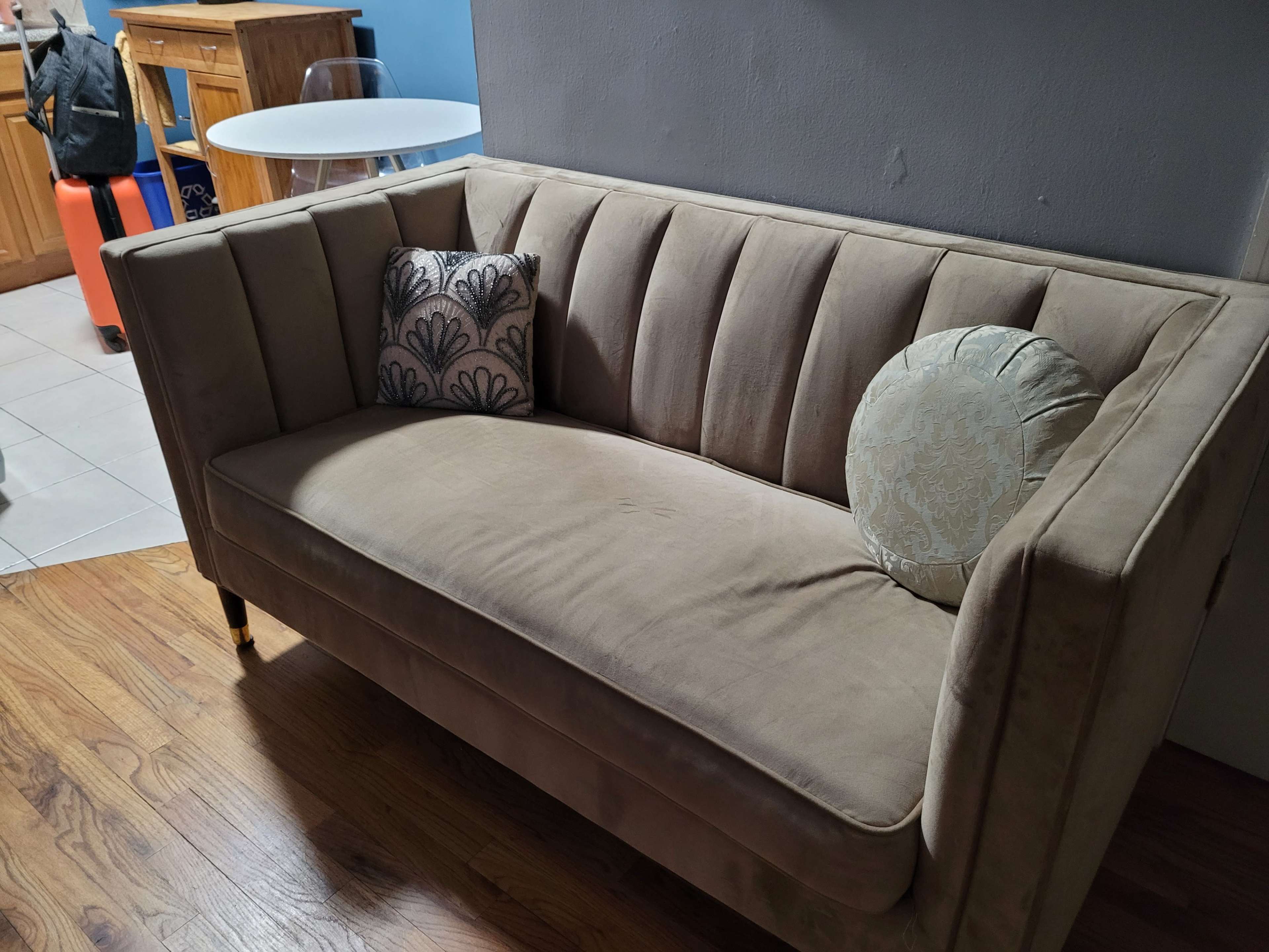A beige couch with vertical tufting and two decorative pillows is positioned in a room with wooden flooring.