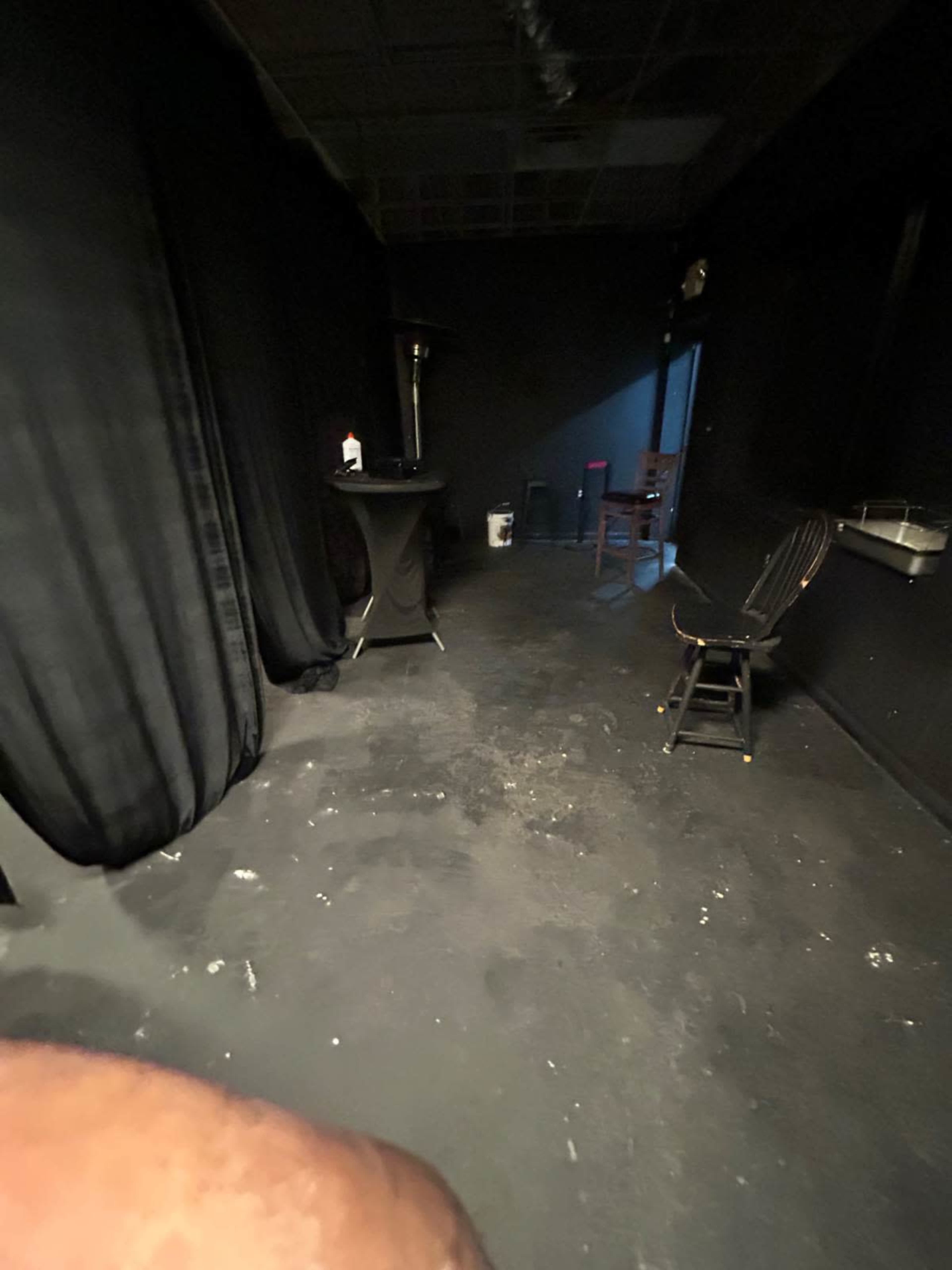 The image shows a dimly lit room with black curtains, a small table, and a wooden chair against the wall.