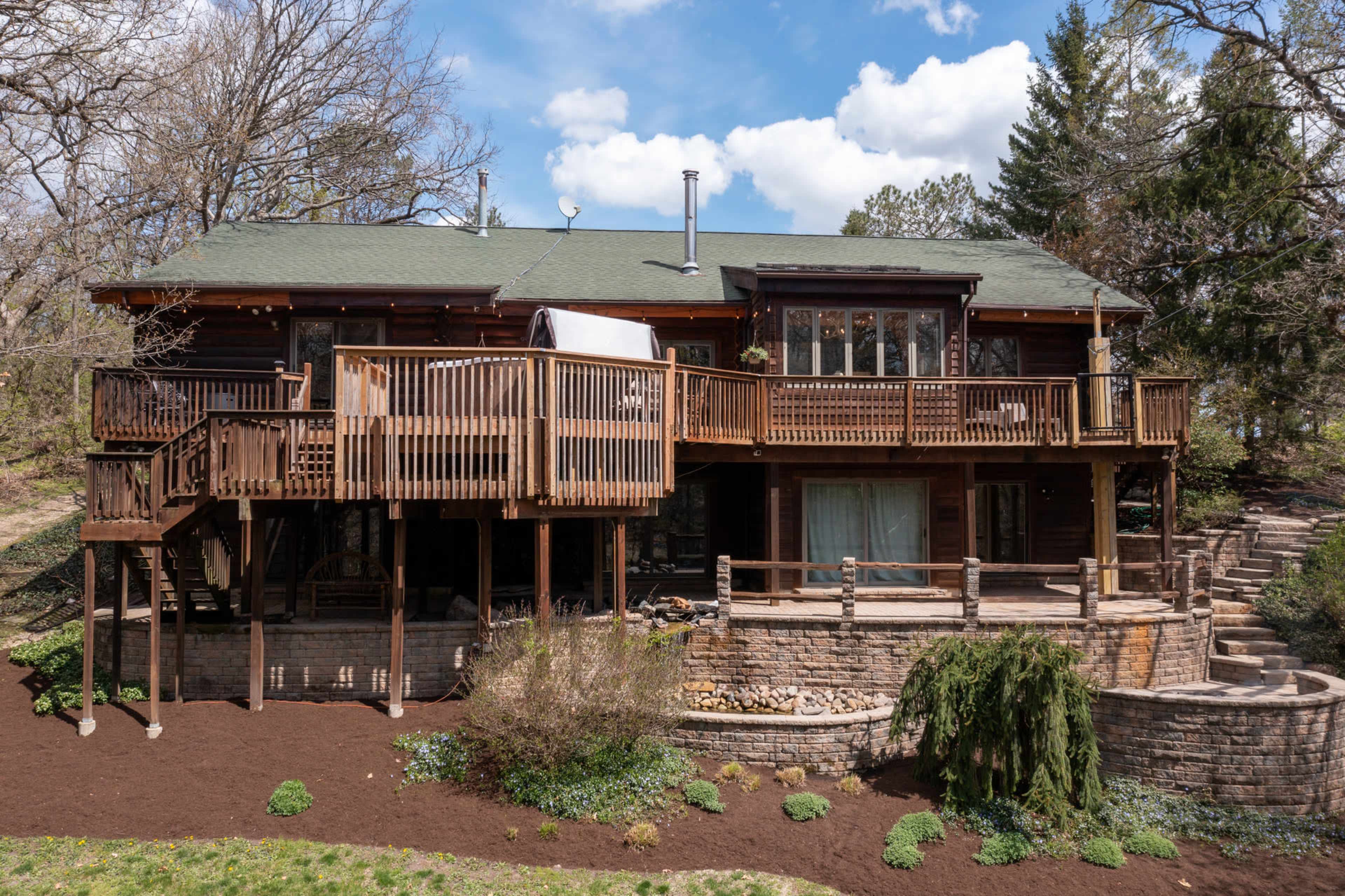 A wooden two-story house with a large deck supported by wooden posts, surrounded by trees and landscaping.