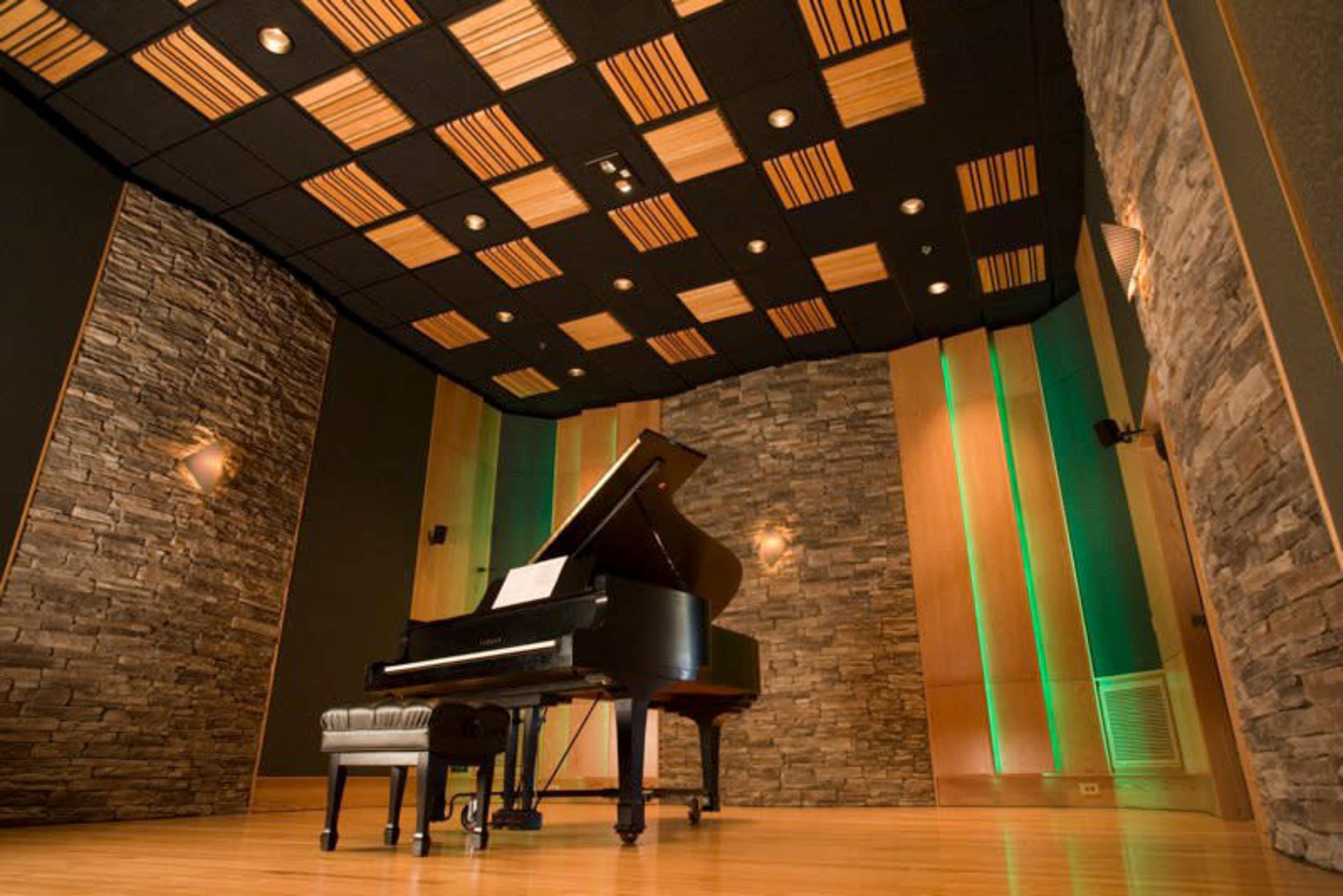 The image shows a grand piano positioned in a soundproof room with stone walls and wooden ceiling tiles.