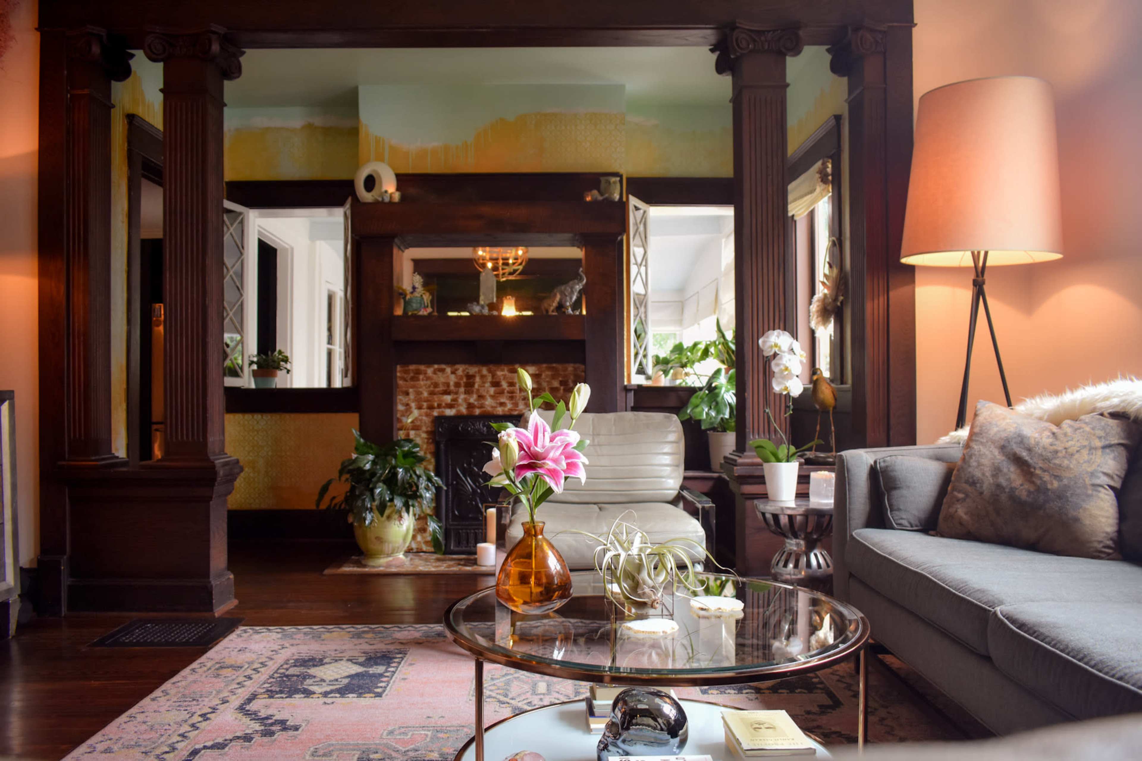 A cozy living room features a gray sofa, a glass coffee table with decorative items, and plants, framed by architectural wooden columns and warm lighting.