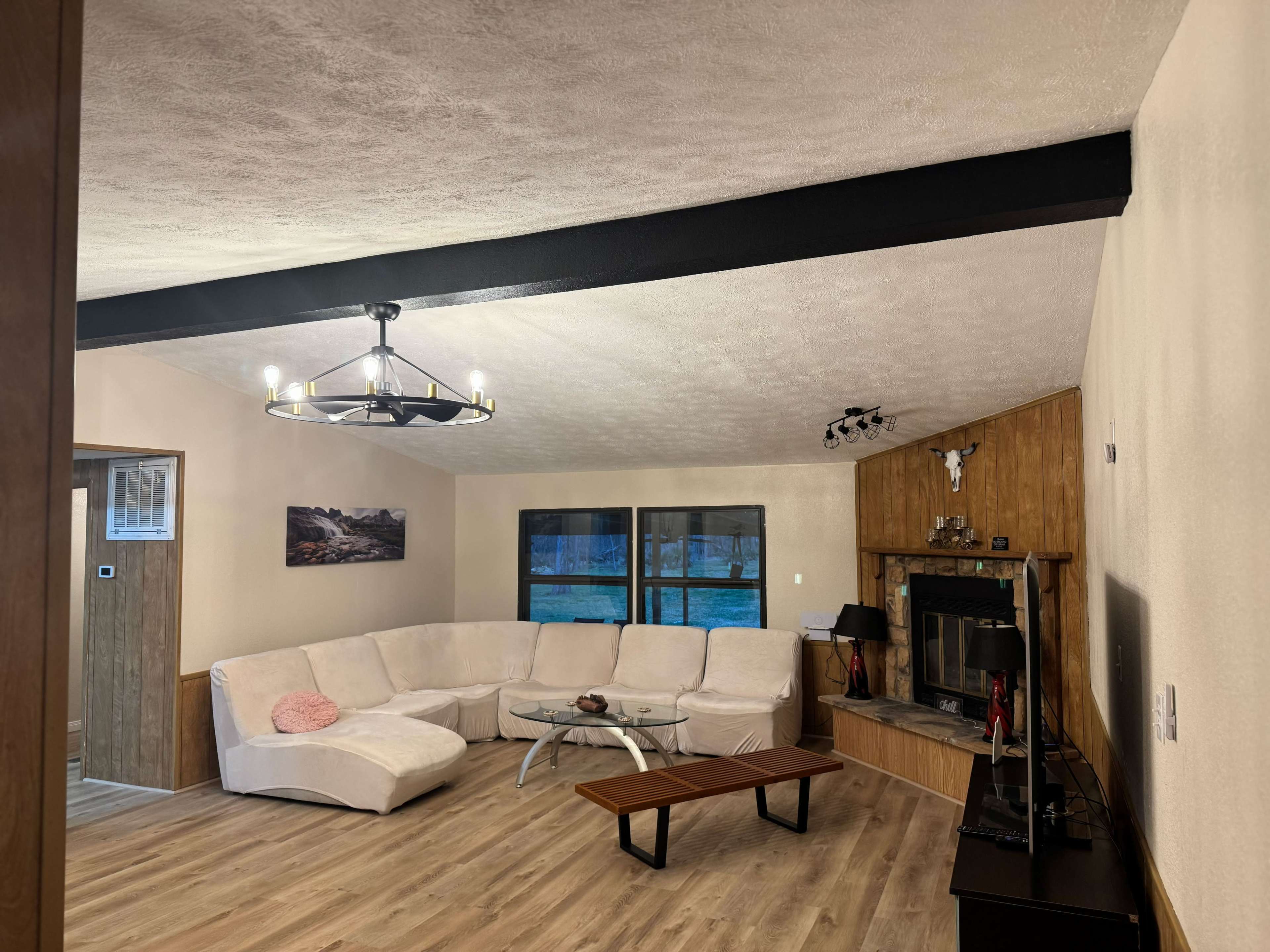 The image shows a living room featuring a large white sectional sofa, a wooden coffee table, a fireplace, and large windows.