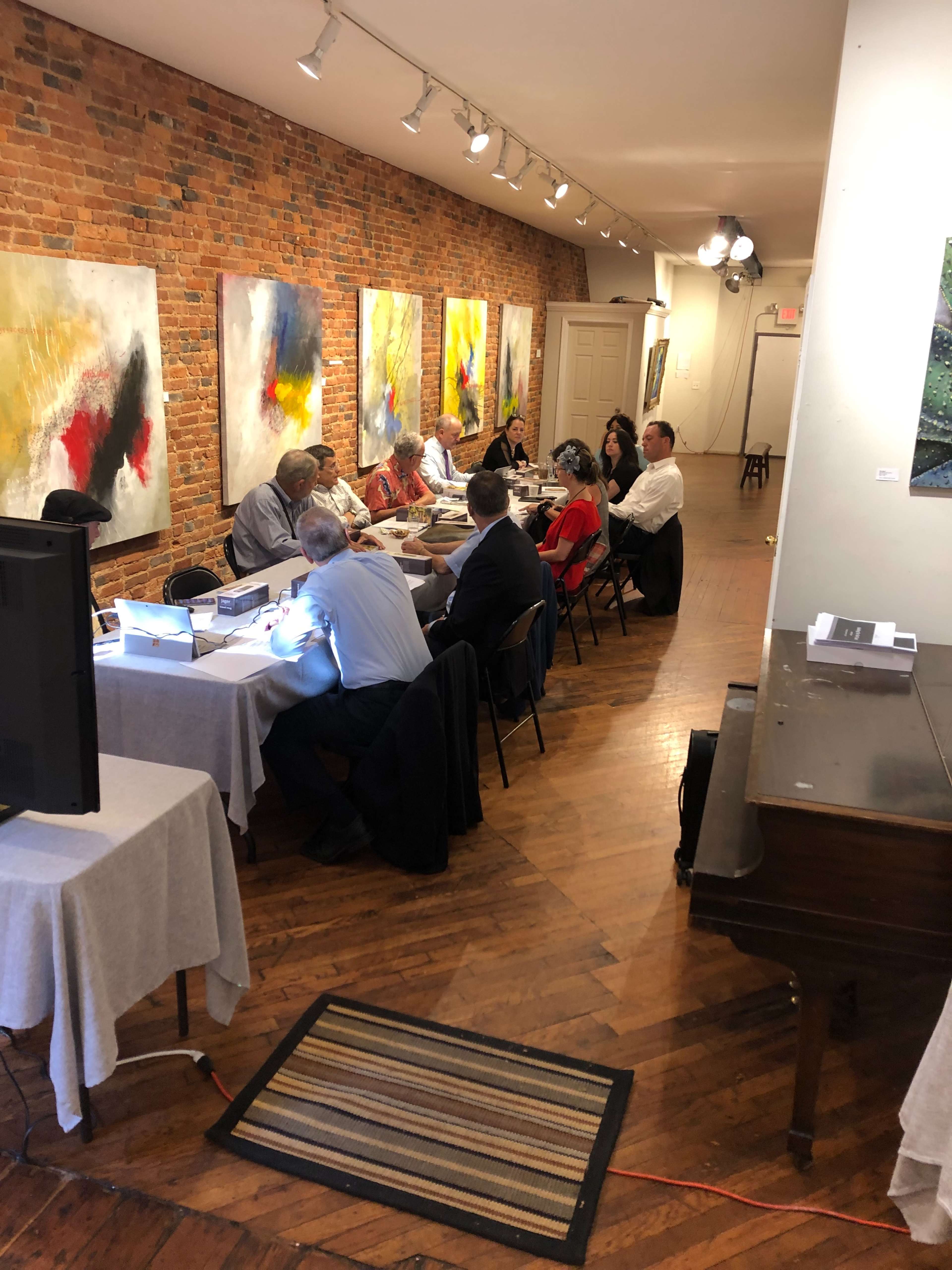 A group of people sits around a long table in an art gallery with colorful abstract paintings on the walls.